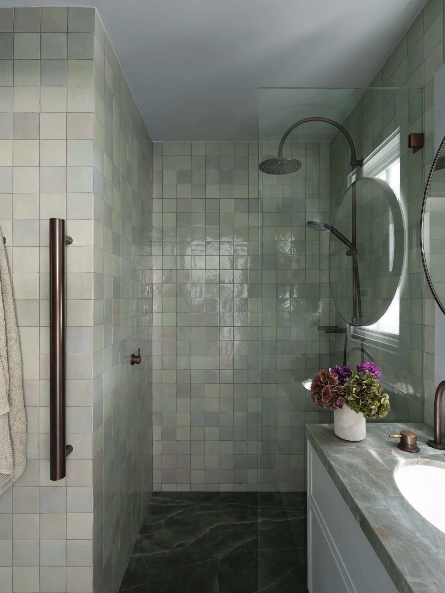 Modern bathroom shower with green tiled walls, rainfall showerhead, handheld shower, large mirror, and a vanity with a marble top. A potted plant with flowers on the vanity and a towel hanging on a hook.