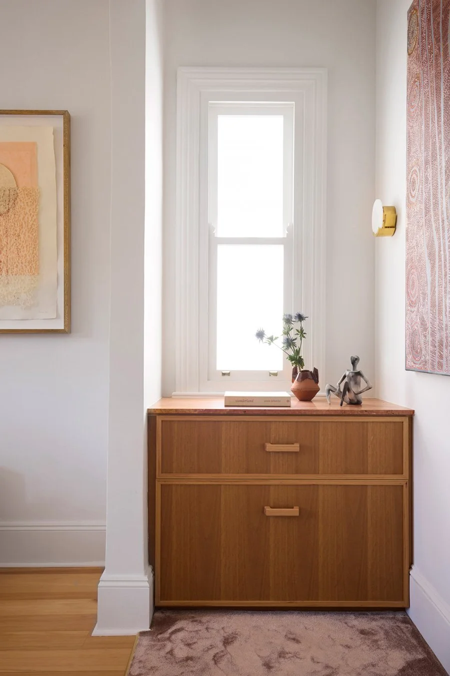 A wooden dresser with two drawers positioned under a window, decorated with a vase of flowers, a book, and a sculpture on top, with abstract artwork on the wall.