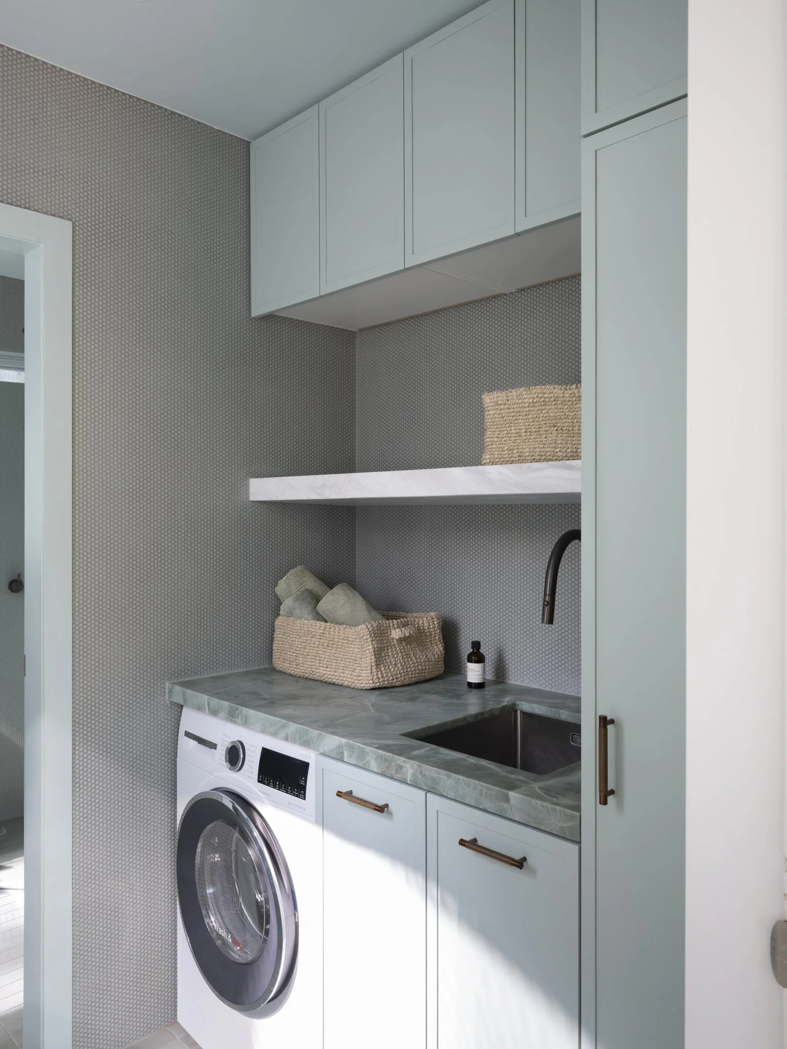 A laundry area featuring a front-loading washing machine, light green cabinets, a marble countertop, a small sink, a woven basket with rolled towels, a brown soap dispenser, and a textured gray wall.