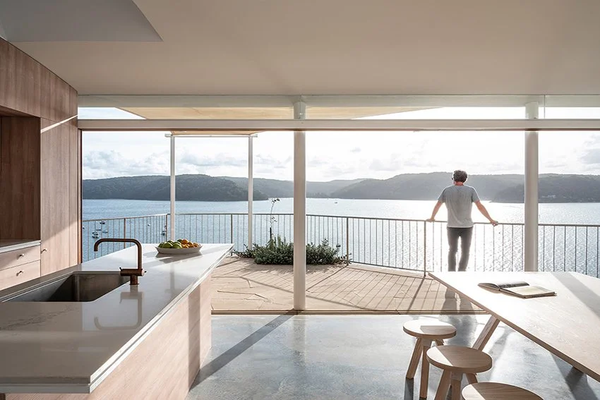 A person standing on a balcony overlooking a large body of water, with mountains in the distance. Inside, a modern kitchen and dining area are visible, featuring a white countertop with a bowl of fruit, a sink with a faucet, and a wooden table with stools. The space is bright and open, with large windows allowing for a view outside.