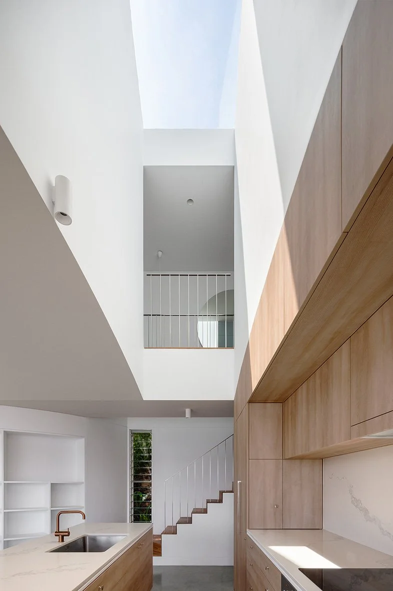 Interior view of a modern home with a kitchen featuring wooden cabinets and a white marble countertop, staircase with white railing, and a mezzanine with a railing visible in the upper part of the image, and skylight letting in natural light.