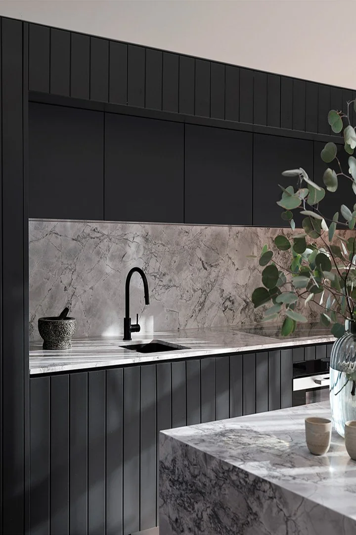 Modern kitchen with black cabinets, marble countertops, and a marble backsplash. Black faucet and a black textured bowl on the counter. Part of a marble kitchen island with a vase of green eucalyptus leaves and small ceramic cups.