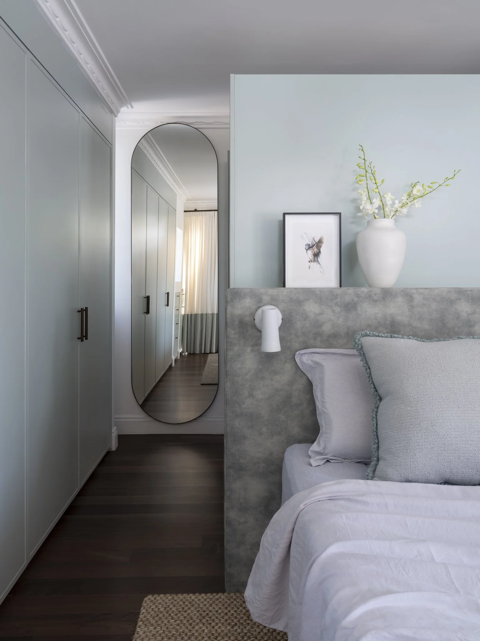 A cozy bedroom corner with a bed with light-colored bedding, a textured throw pillow, and a blanket. A grey upholstered headboard is visible. On the wall above the bed, there is a framed picture of a bird. A white vase with white flowers is placed on a ledge or shelf next to the picture. To the left, there is a tall wardrobe with black handles, and a large oval mirror reflecting part of the room and a doorway with curtains. The room has dark wood flooring and a small woven rug near the bed.