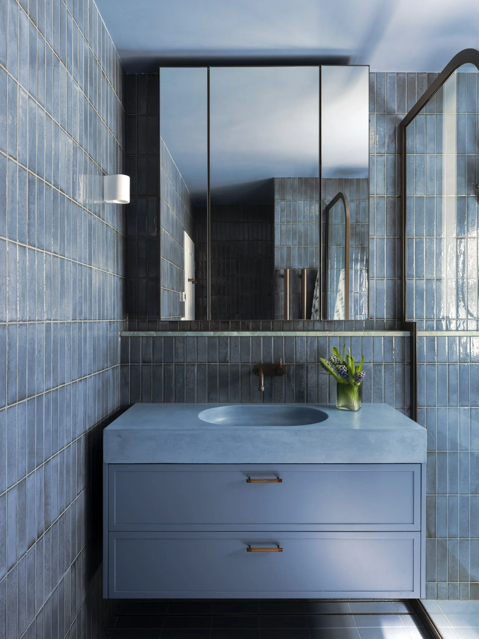 Modern bathroom with blue tiled walls, a white vanity with a round sink, and a large mirror reflecting the sky and a building. A small potted plant is on the vanity.