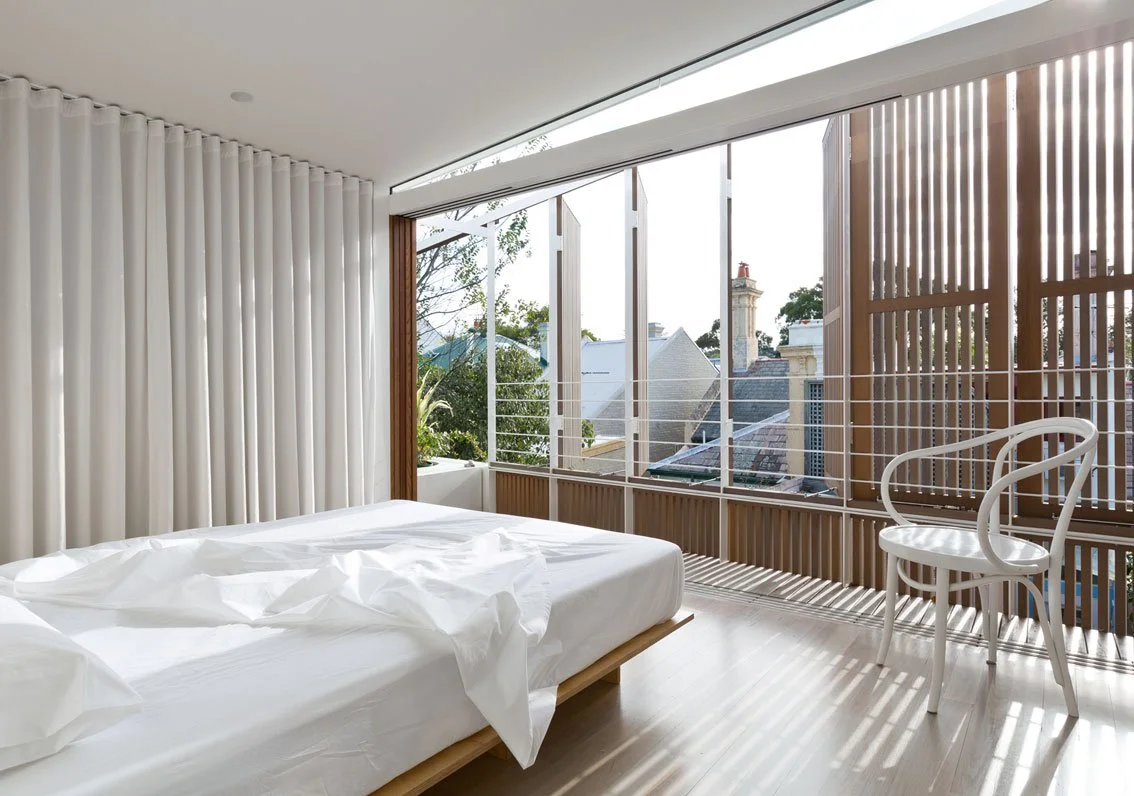 A modern bedroom with a large bed with white bedding, white curtains, and a white chair. The room has large windows with vertical wooden slat blinds, letting in natural light and offering a view of rooftops and trees outside.