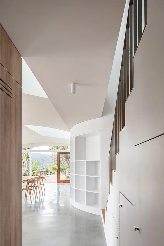 Modern interior with curved white shelving, wooden staircase, and large window with outdoor view.