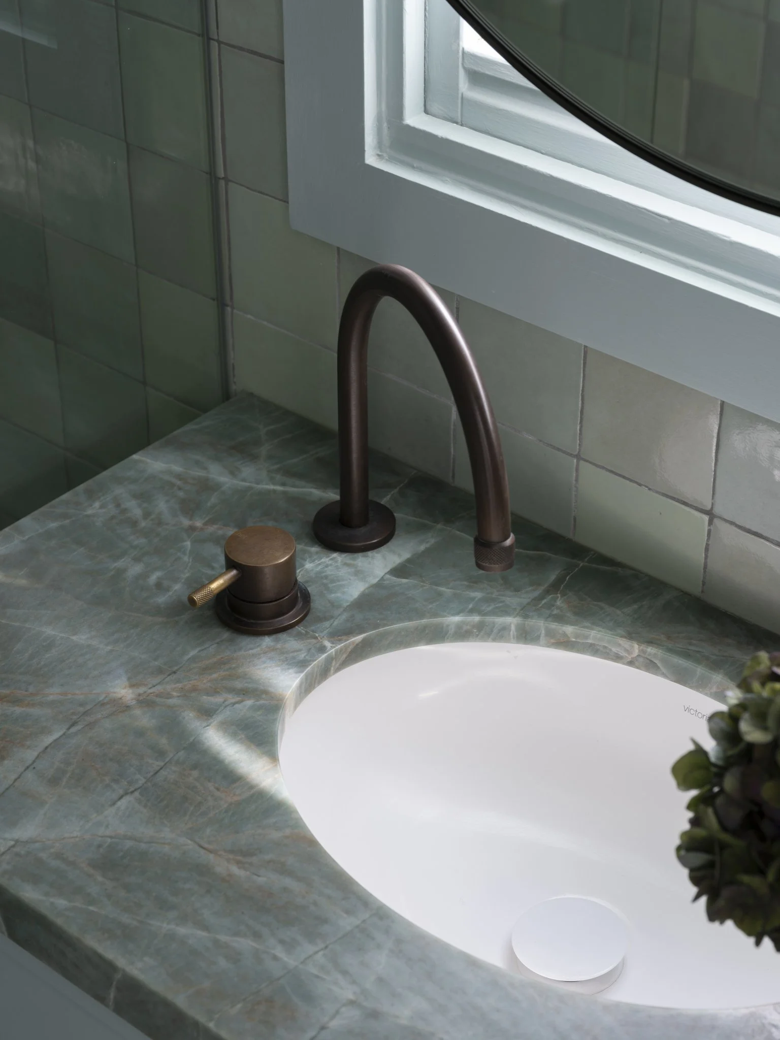 Close-up of a bathroom sink with dark bronze fixtures, a green marble countertop, and a window with a white frame, letting in natural light.