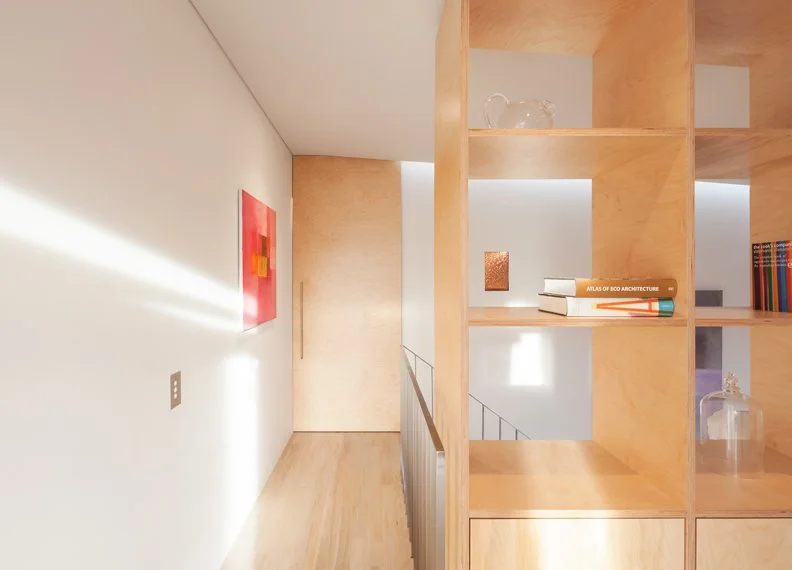 Interior view of a modern home with a wooden bookshelf, a staircase, and a white wall with artwork and lighting.