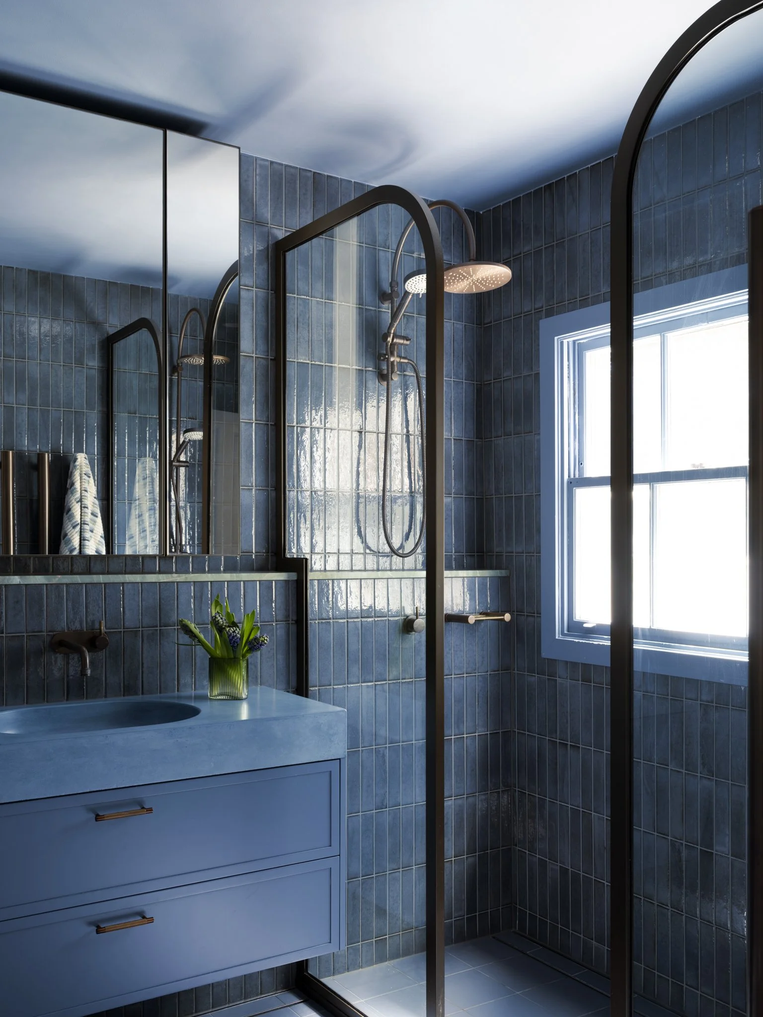 Shower with black frame glass doors, blue tiled walls, and a ceiling-mounted round rain shower head, next to a window with a white frame, near a blue vanity with a vase of purple flowers.