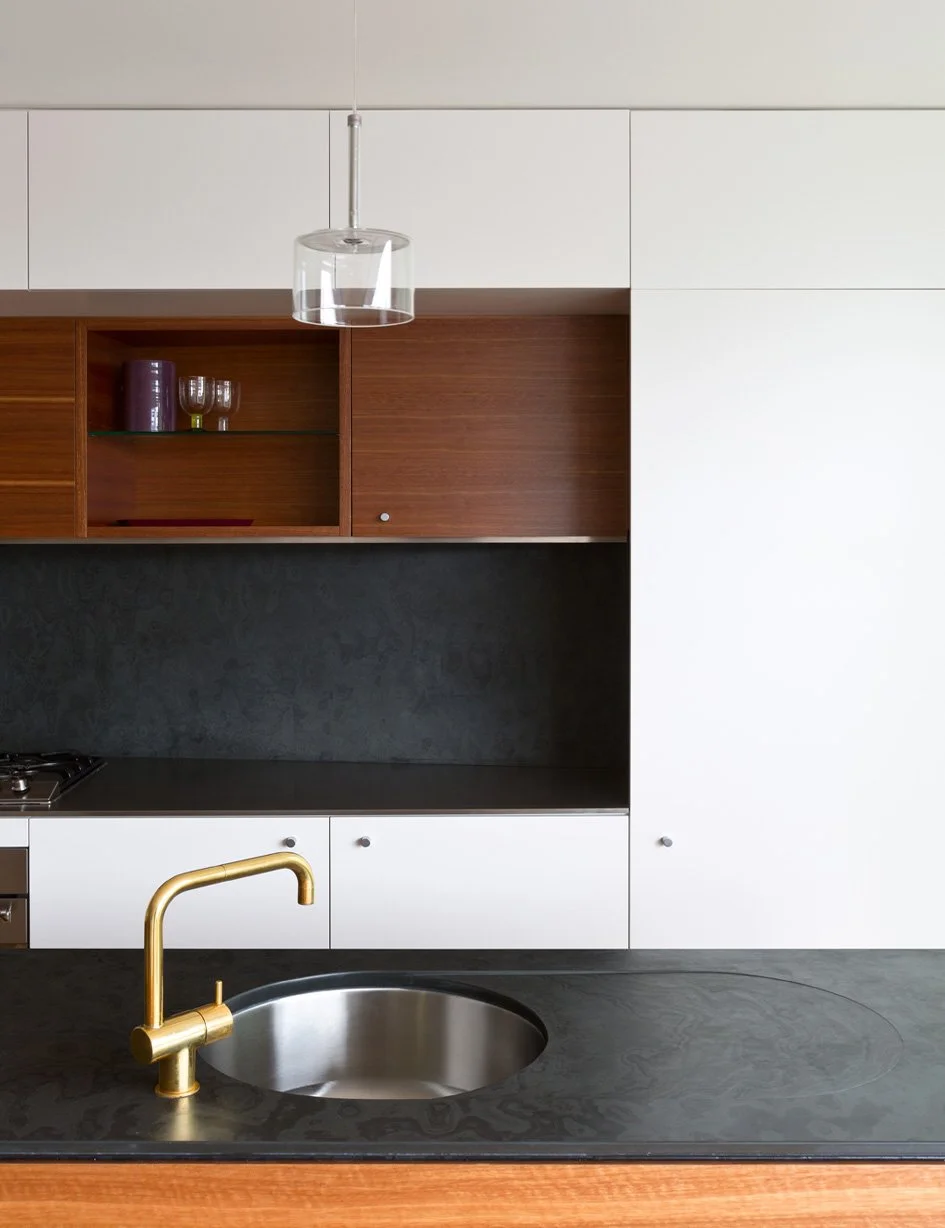 Modern kitchen with black countertop, brass faucet, white cabinets, wooden open shelves, black backsplash, and transparent pendant light.