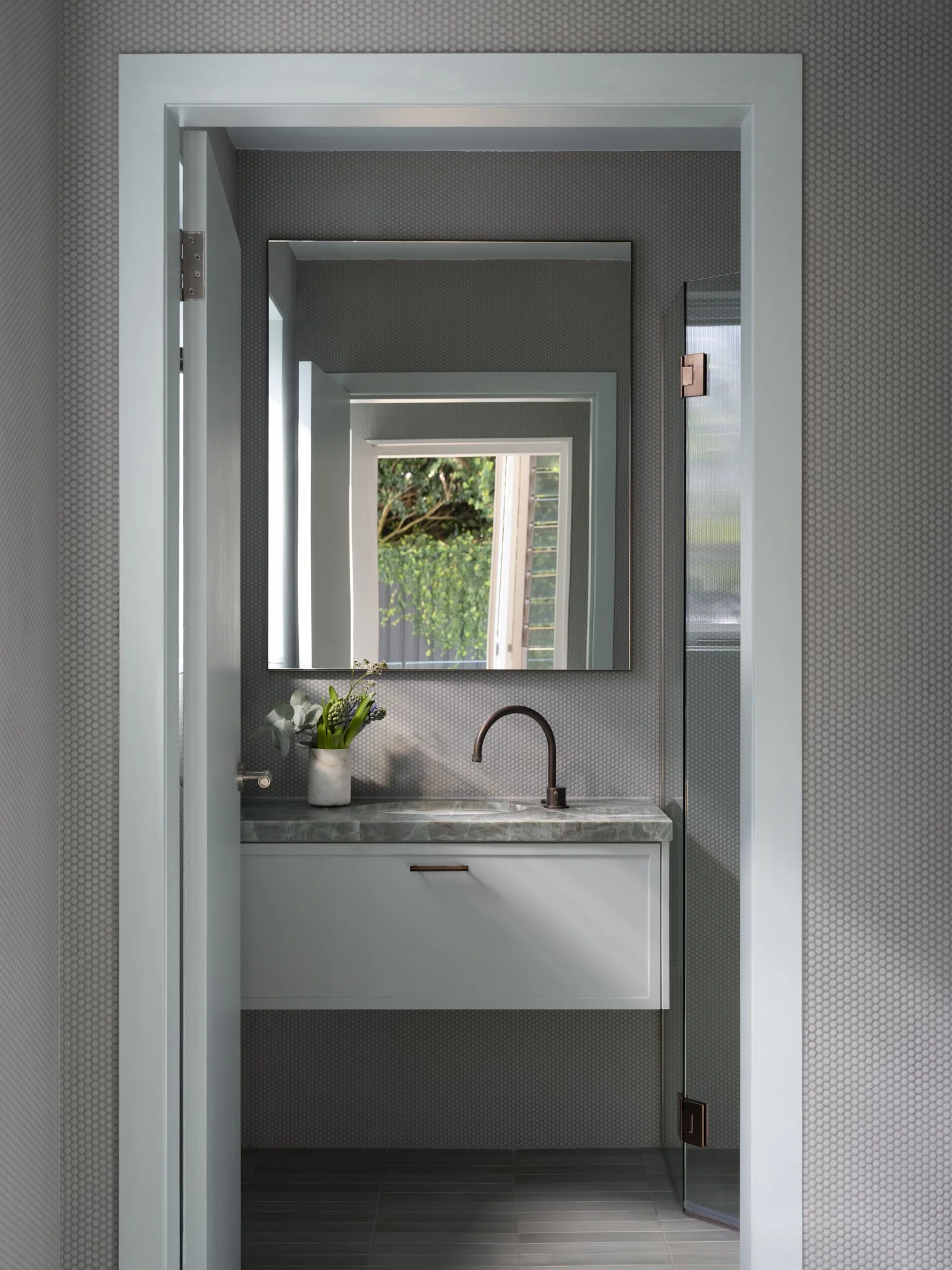 A modern bathroom sink area with a marble countertop, a vase of flowers, a faucet, a mirror, and a window showing greenery outside.