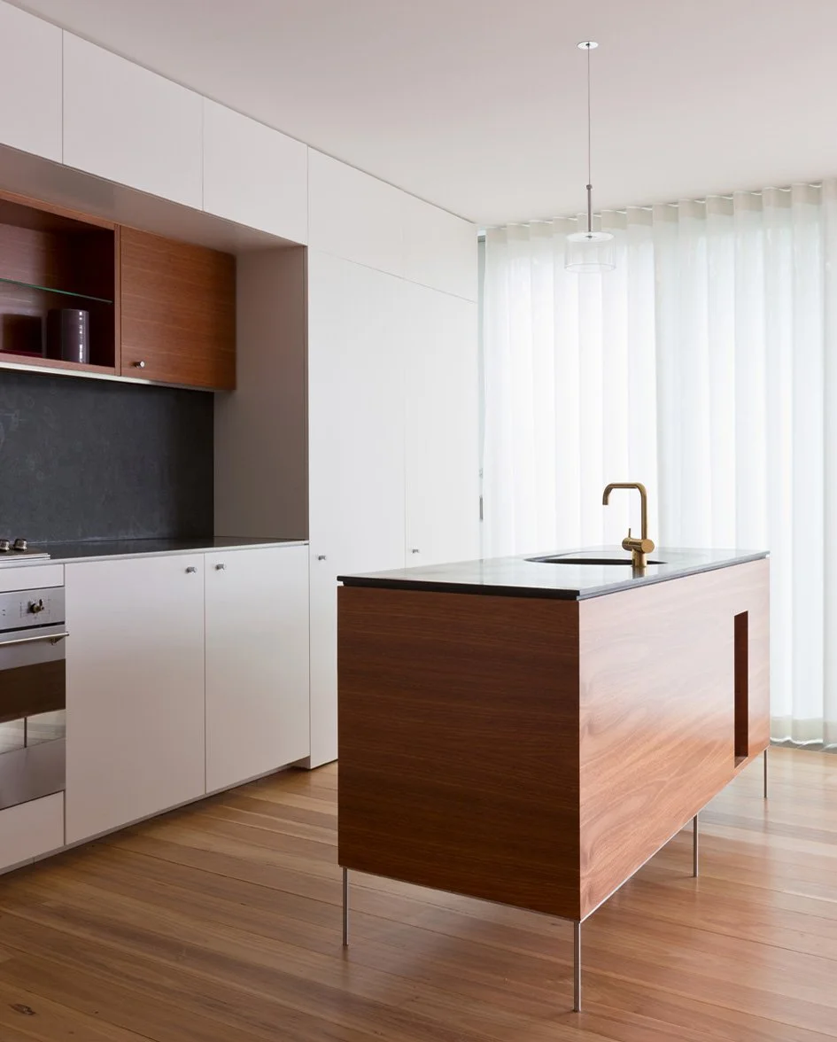 Modern kitchen with white cabinets, a wooden island with a black countertop, and a brass faucet, set against sheer white curtains and wooden floors.