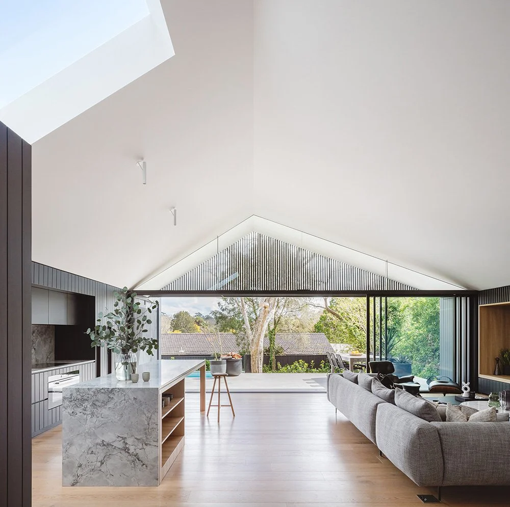 Modern living room with a high, sloped ceiling, large glass doors opening to a backyard, and contemporary furniture including a gray sofa and black lounge chairs.