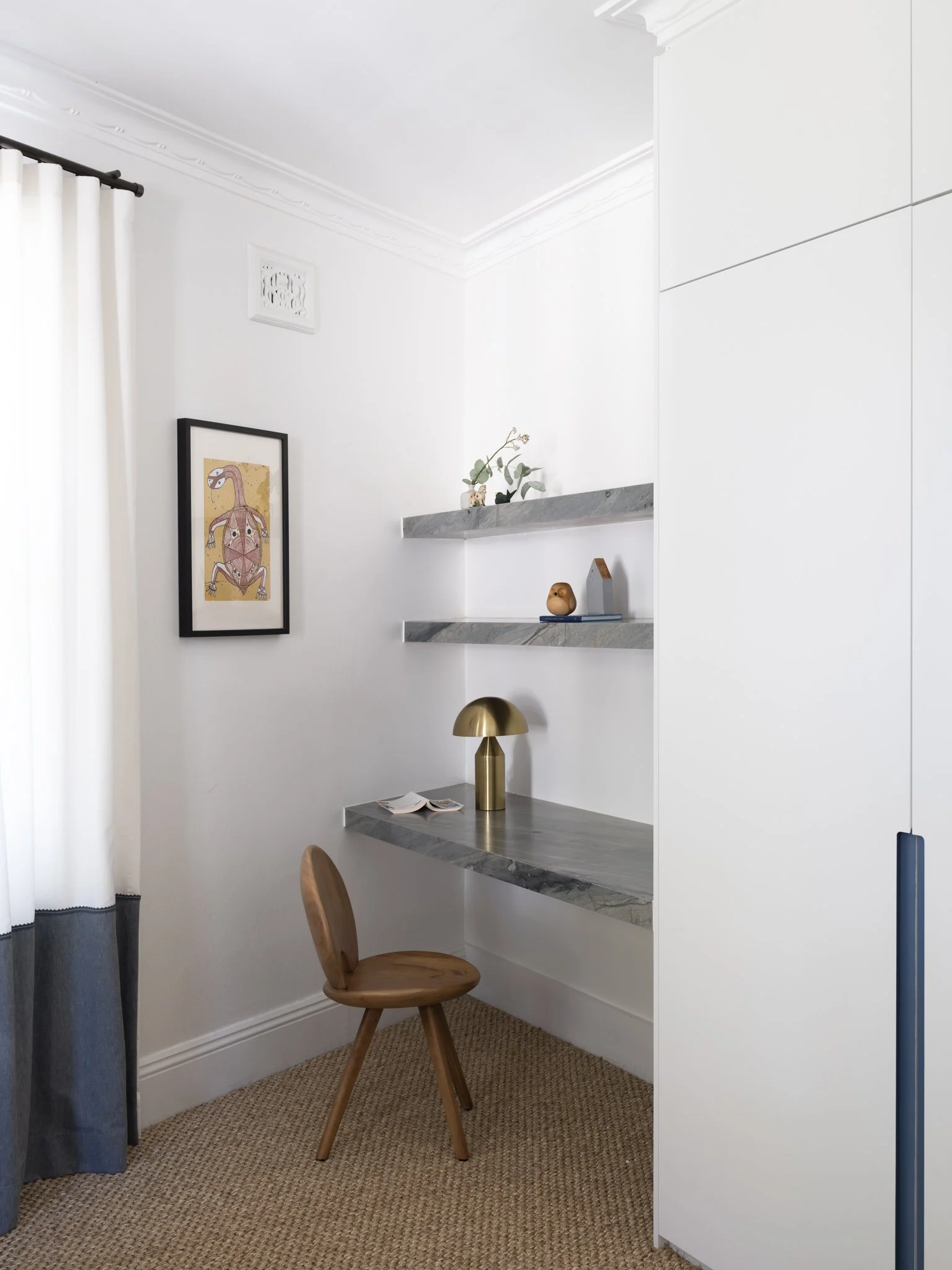 Corner of a minimalist home office with white walls, a wooden chair, floating marble shelves decorated with books, a small plant, and decorative objects, and a gold table lamp.