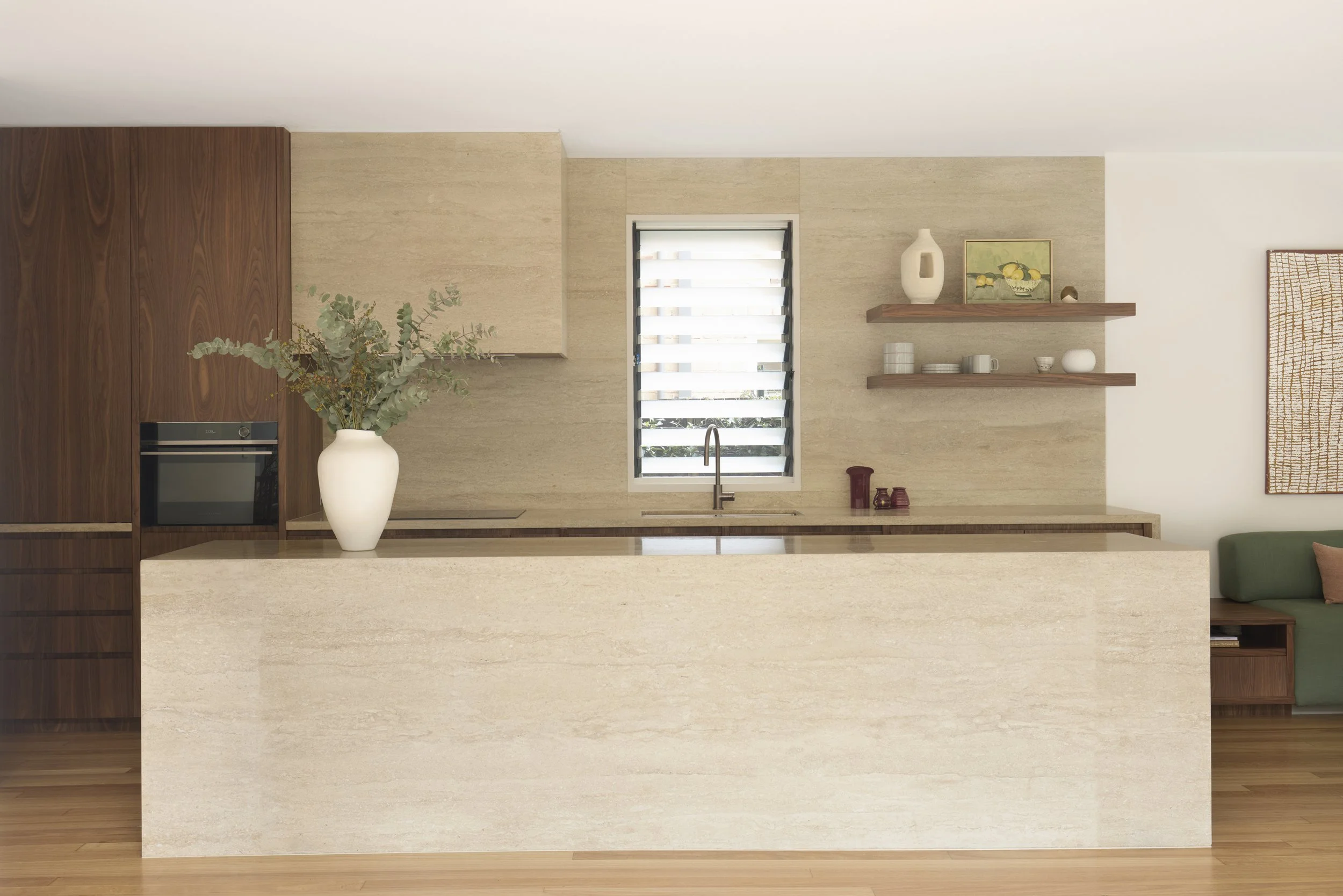 Modern kitchen with beige and wood finishes, large window with blinds, decorated with vases, artwork, and shelves.