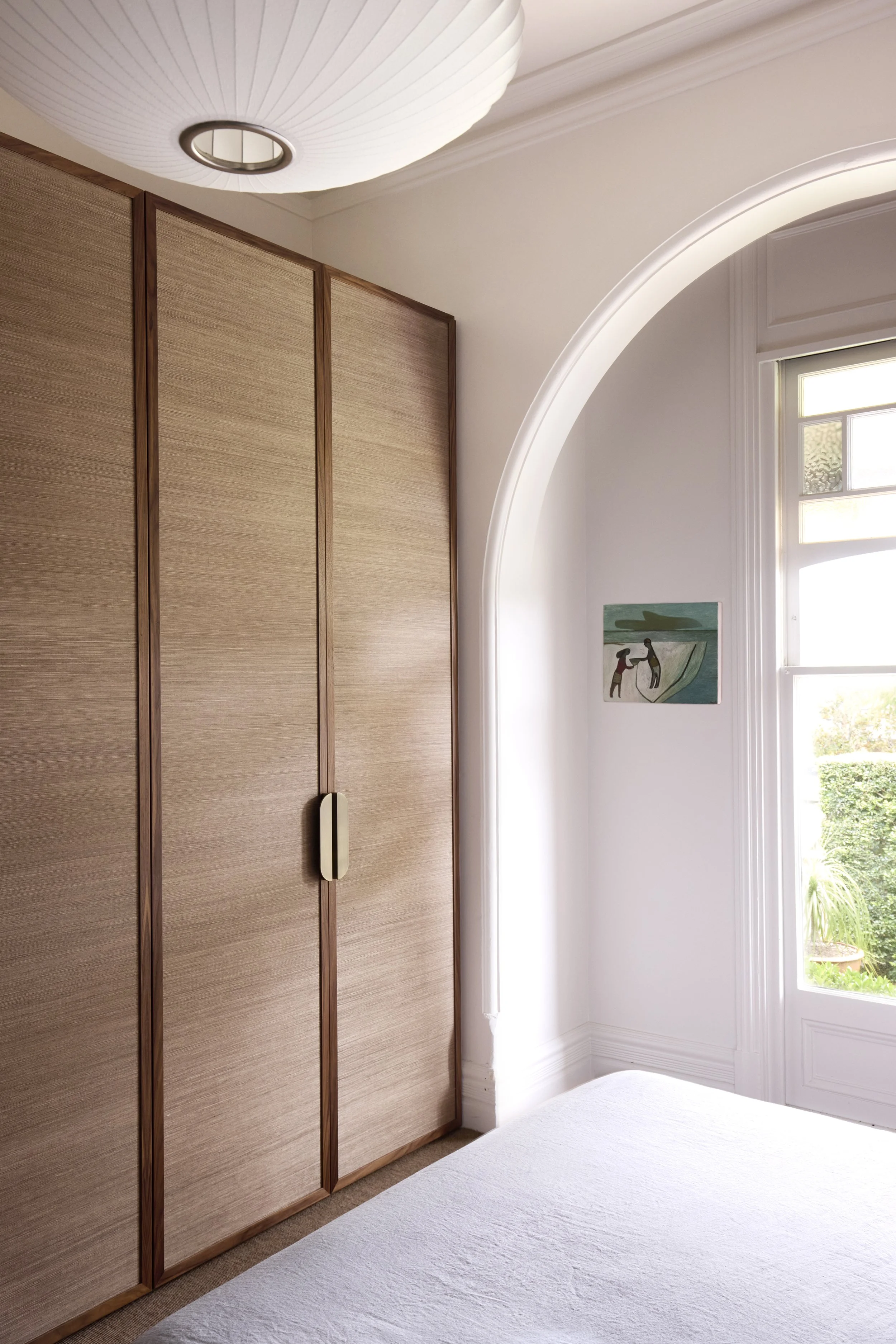 A bedroom with a wooden wardrobe, white wall, artwork, and a window with sunlight.