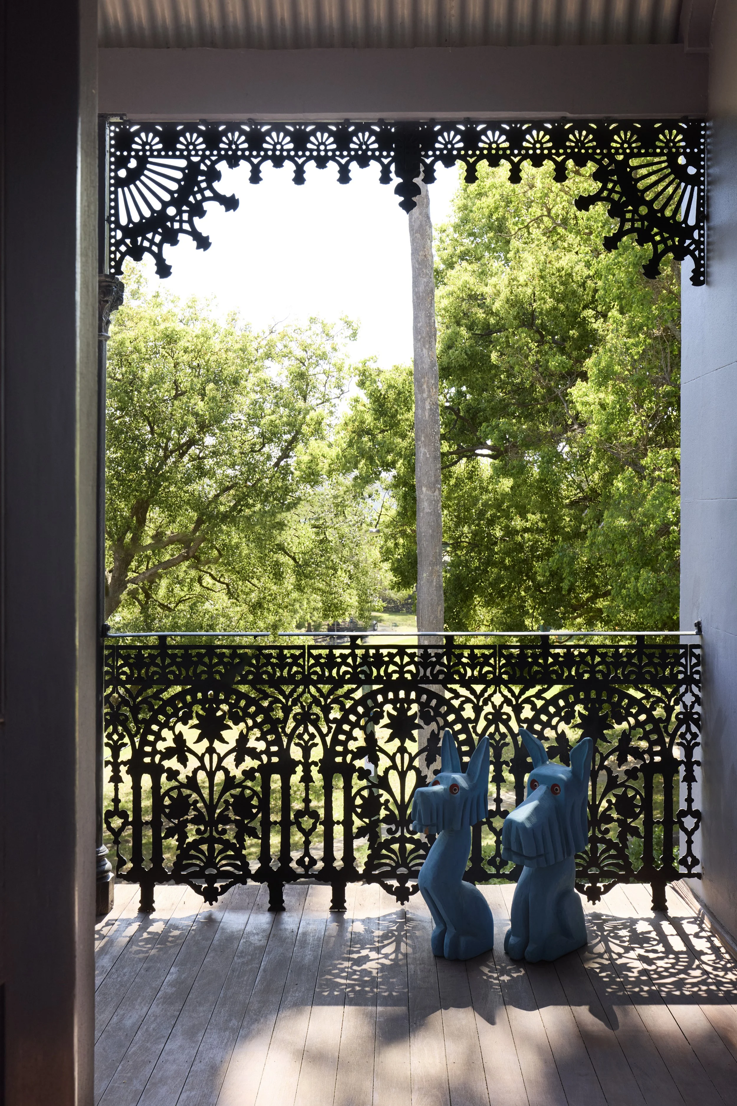 View of a porch with ornate black metal railing and two blue dragon sculptures on the floor, overlooking trees with green foliage under bright sunlight.
