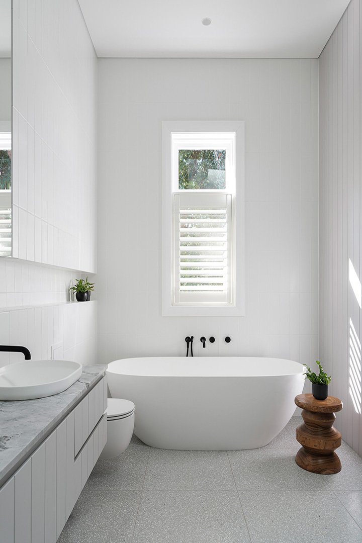 Modern white bathroom with a bathtub, toilet, and a vanity with a vessel sink. Natural light through a window with white shutters. Small potted plants on a wooden side table and the vanity.