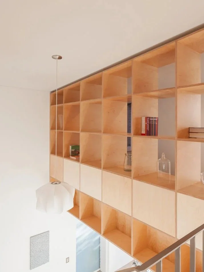 Wooden grid-style bookshelf with empty shelves and a few books and decorative items, hanging light fixture, white wall, and staircase railing in the foreground.