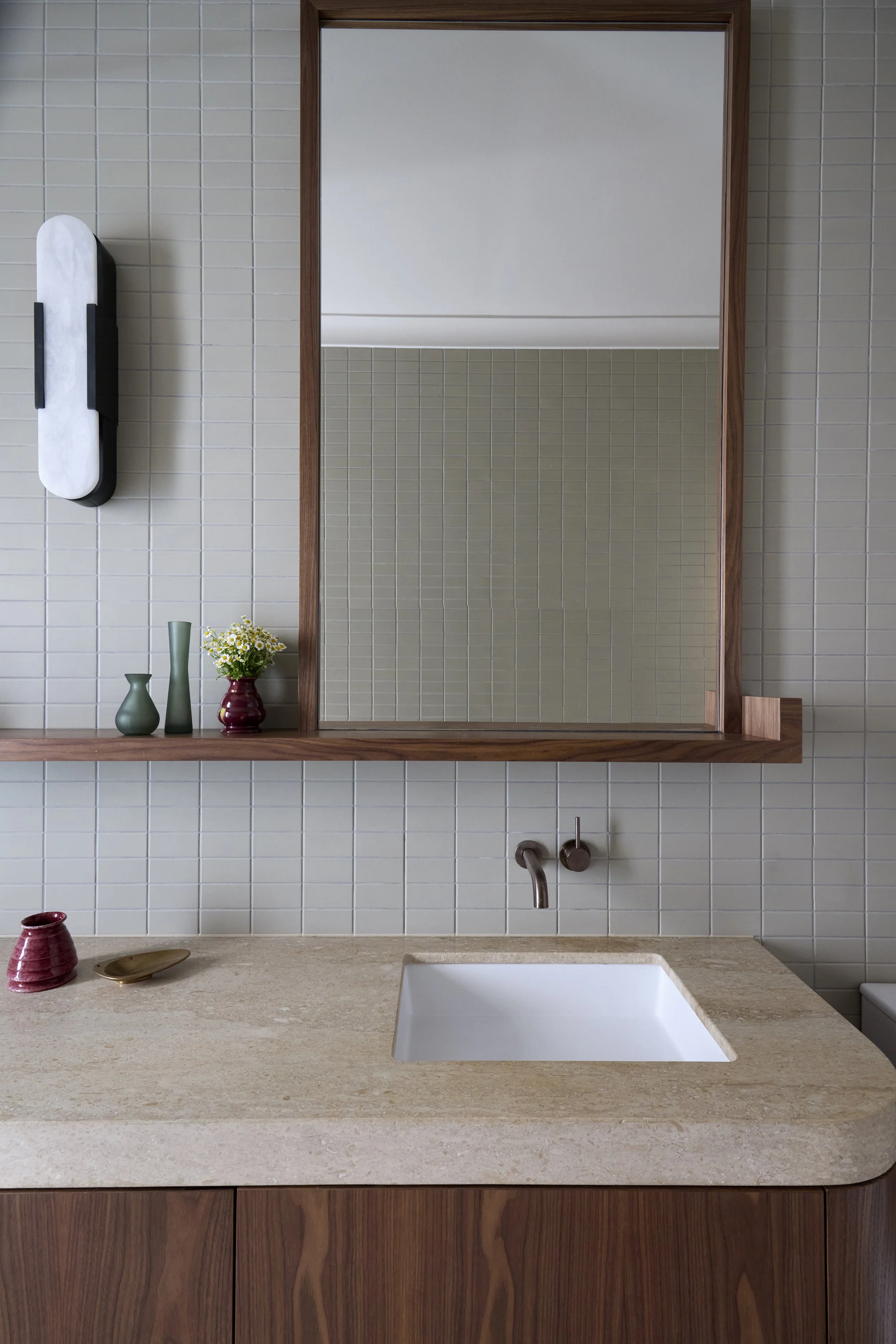 A modern kitchen with beige stone countertop, white sink, and pastel green and maroon vases on a wooden shelf. Large mirror on beige tiled wall reflects ceiling lights and part of the room.