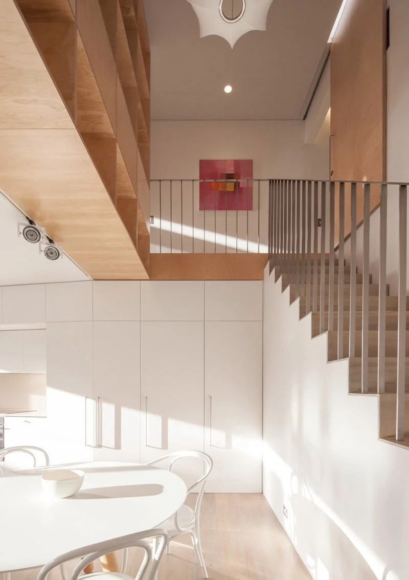 Interior view of a modern home with a white dining table and chairs, wooden staircase with vertical railings, white cabinetry, and a pink abstract painting on upper wall.