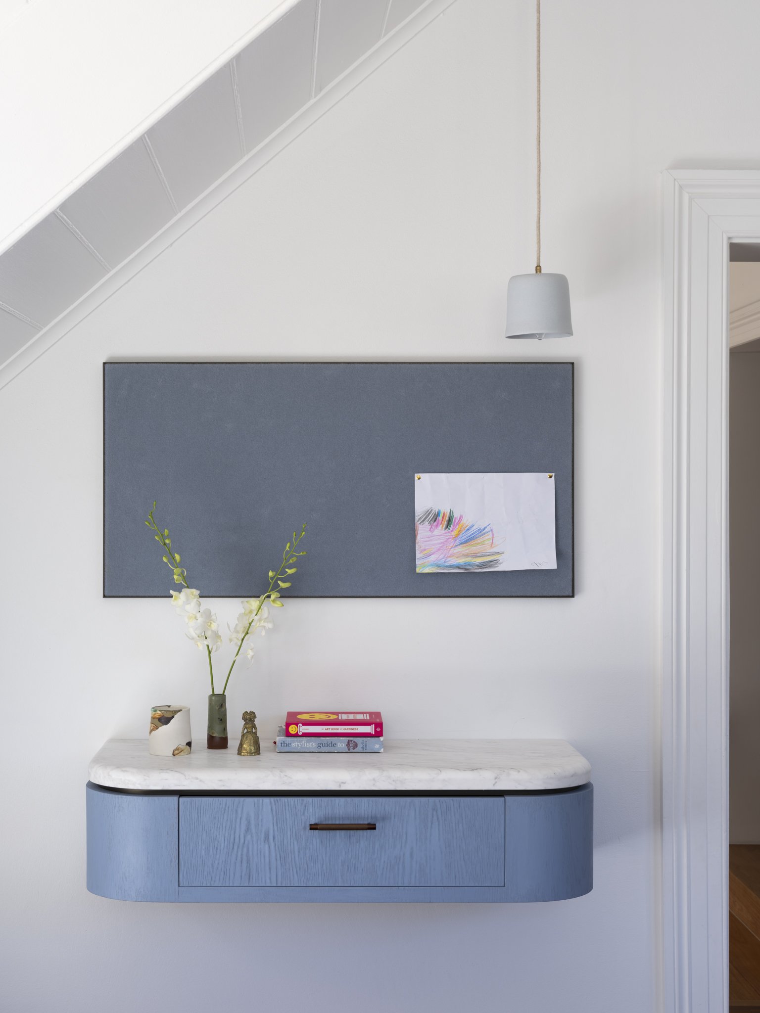 View of a decorative wall shelf with a blue finish, mounted on a white wall. The shelf holds a white marble vase with white flowers and green foliage, a smaller brown vase, and a golden figurine. Below, on a white surface, are three books stacked, titled 'The Stylist's Guide to,' 'Art Book of Happiness,' and another partially visible. The background includes a black bulletin board with a colorful drawing and a framed doorway to the right. A white pendant light hangs from the ceiling on a beige cord.