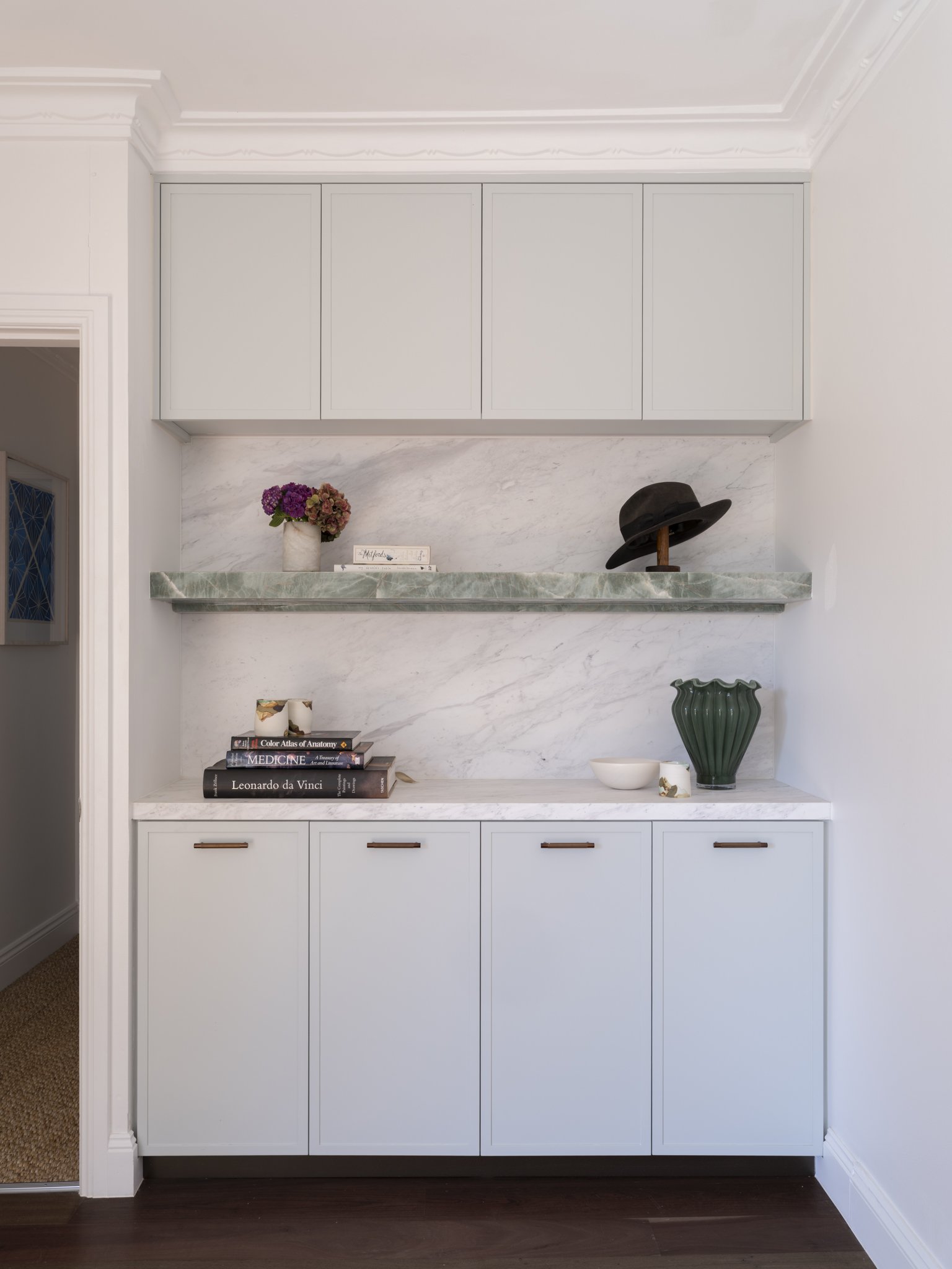 A white built-in cabinet with four doors mounted on the wall above a marble countertop. On the countertop, there is a vase with purple and pink flowers, a stack of books, a white bowl, and a large black hat displayed on a wooden stand. There is also a green decorative vase and a small white item on the lower shelf. The room has wooden flooring and crown molding along the ceiling.
