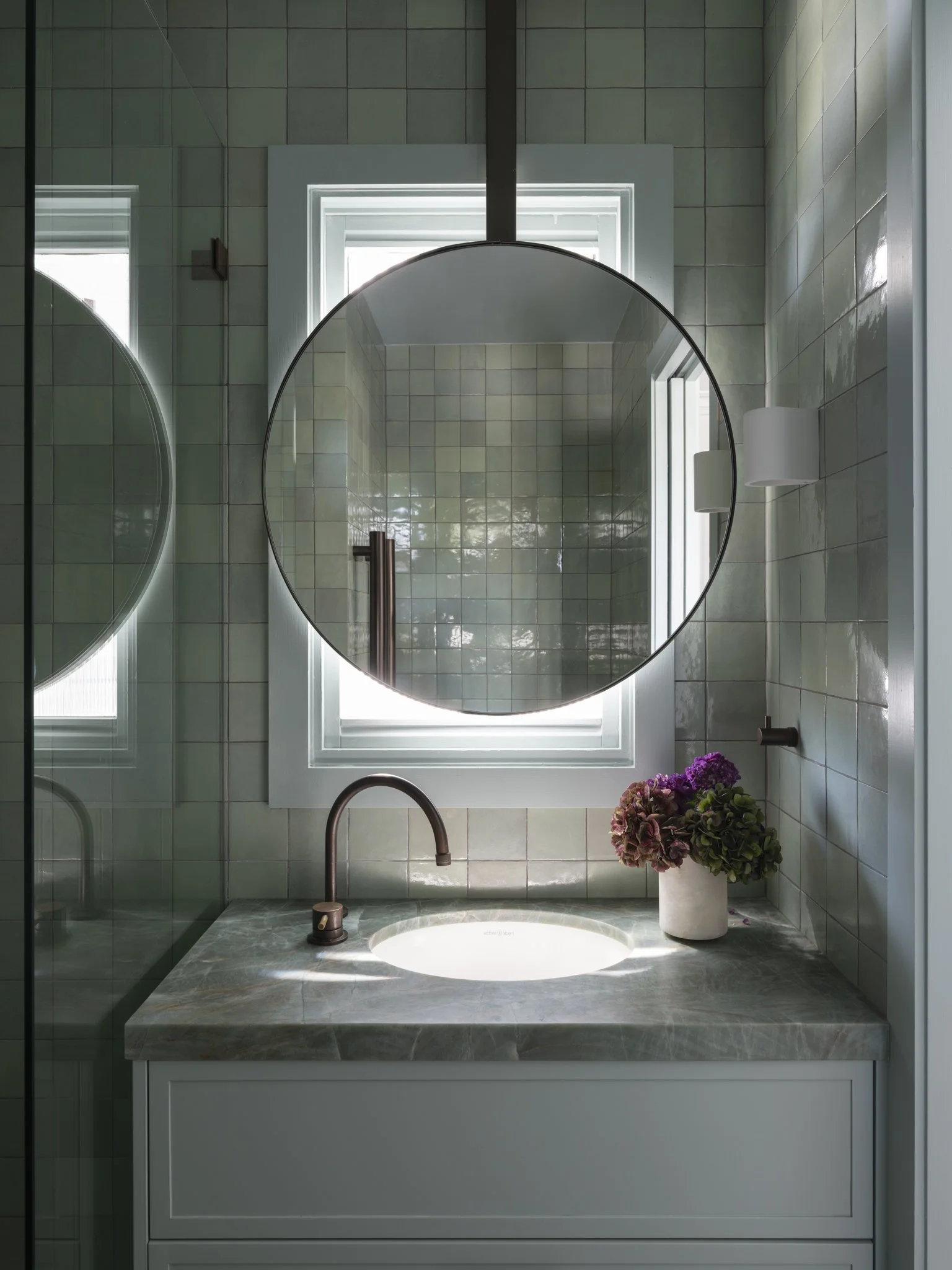 Bathroom vanity with a round mirror, a sink, a bronze faucet, a window above, a white flowerpot with purple hydrangeas, and cream tiles on the wall.