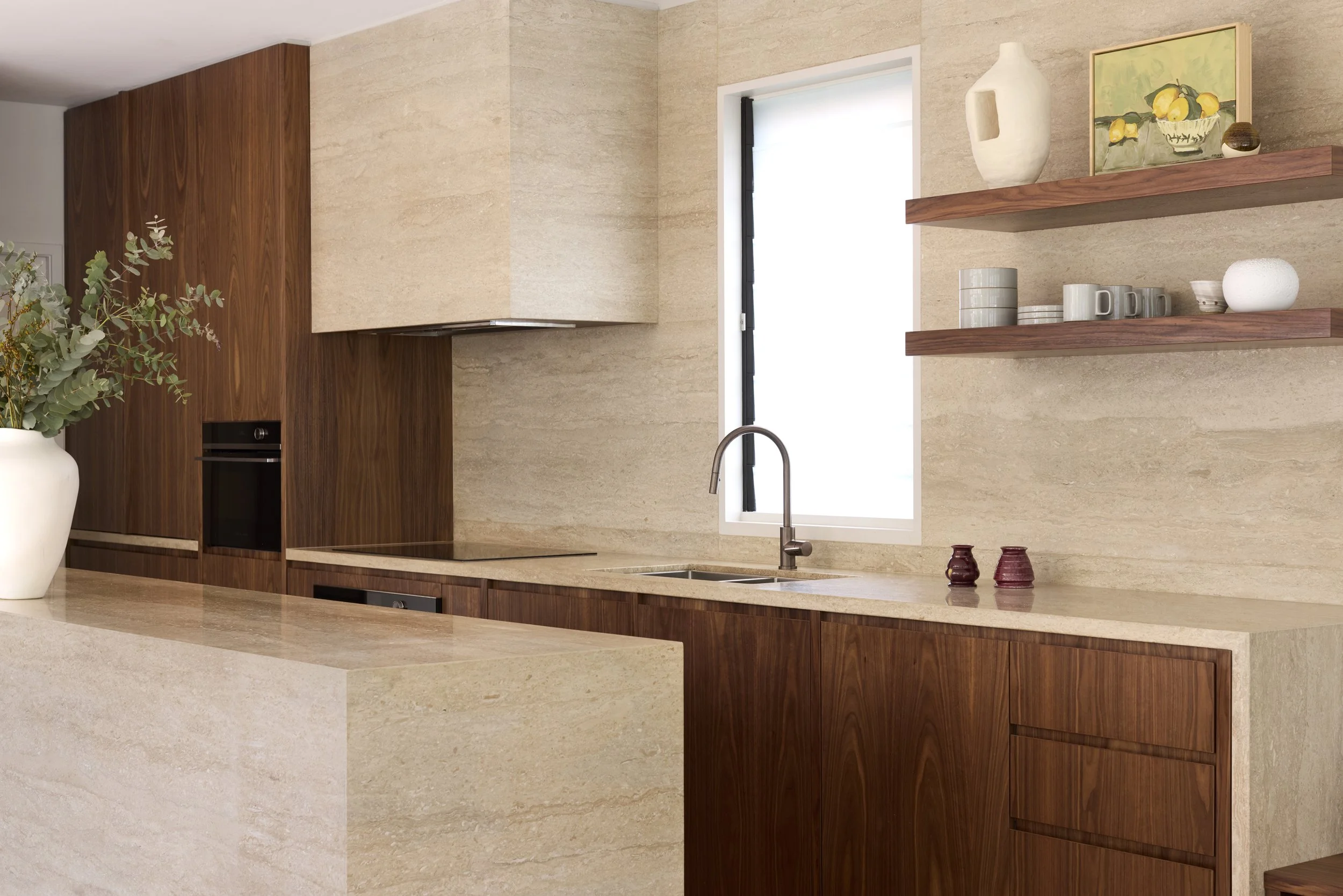 Modern kitchen with beige stone walls, wooden cabinetry, a window, and open shelves displaying ceramic dishes and artwork.