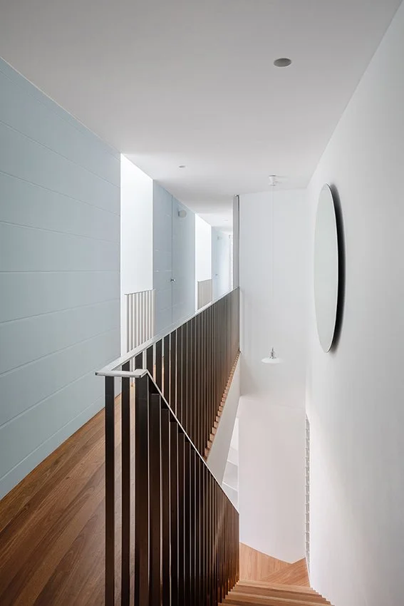 Interior view of a narrow hallway with a wooden staircase and black metal railing, white walls, and a round mirror on the wall.