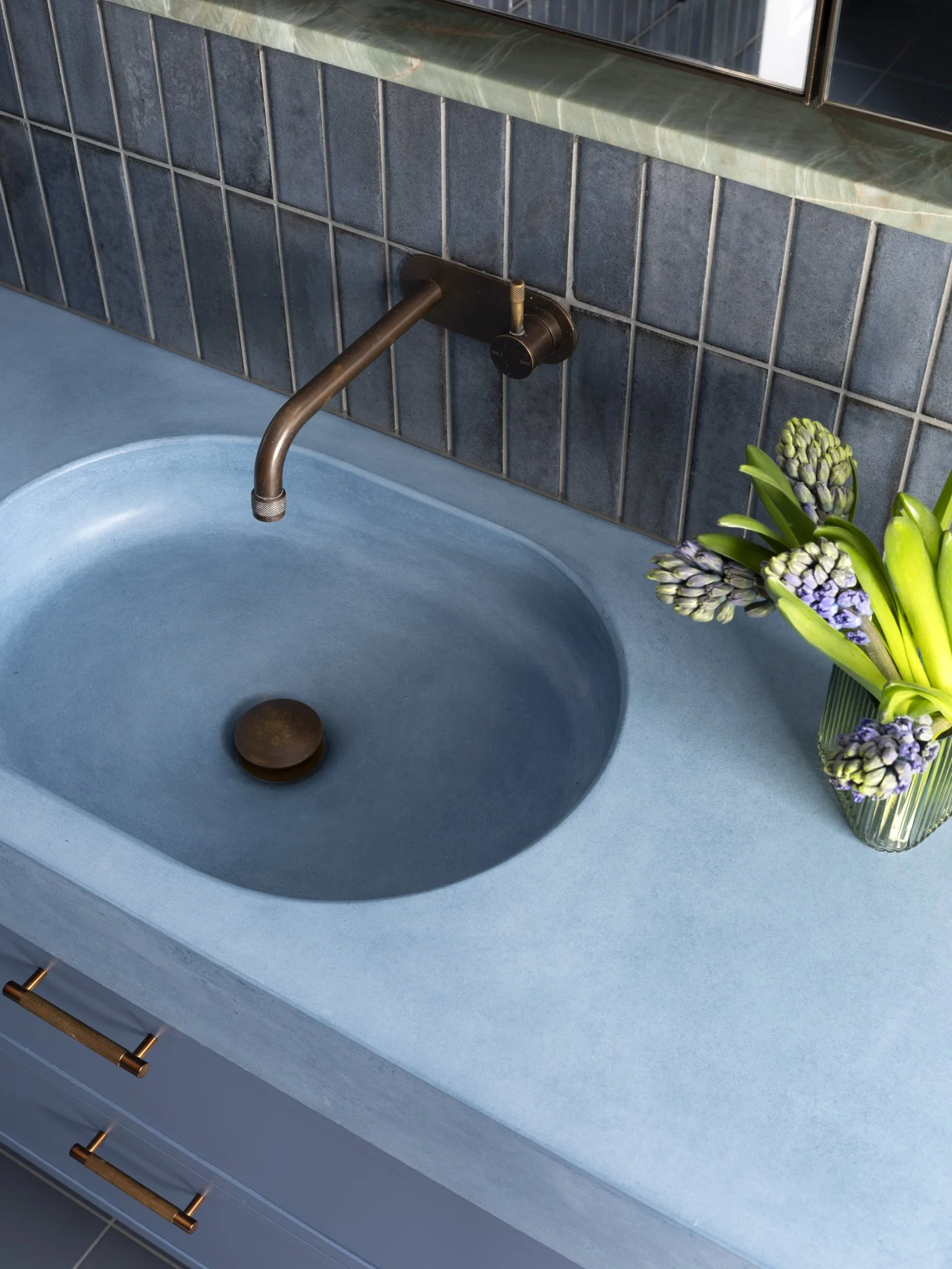 A blue kitchen sink with a bronze faucet, a brown soap dish, and a vase of purple and green flowers on the countertop. The background features a tiled backsplash and a mirror above the sink.