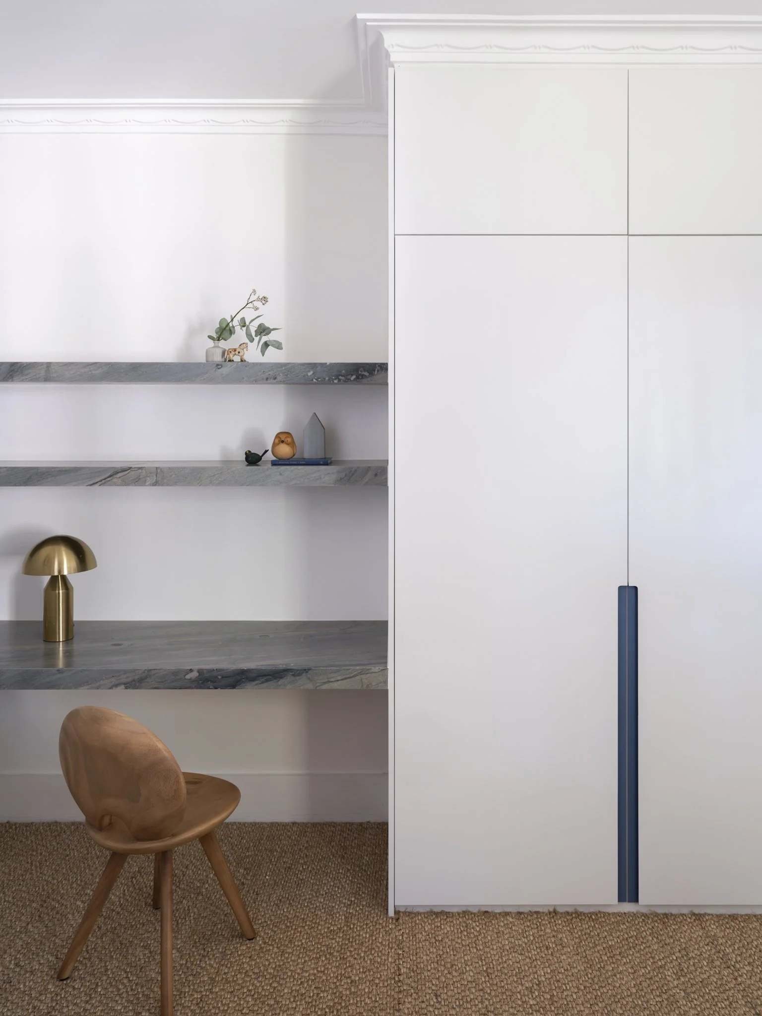 A modern interior space with a wooden chair, built-in shelves with decorative items, a metallic table lamp, and a white cabinet, with a woven carpet on the floor.