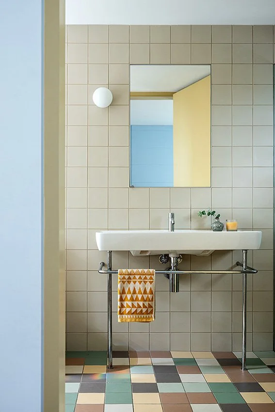 A bathroom with beige tiled wall, a rectangular mirror, a white sink with metal pipes, a yellow patterned towel hanging on a towel bar, a small potted plant, and a candle on the sink, with a colorful tiled floor.
