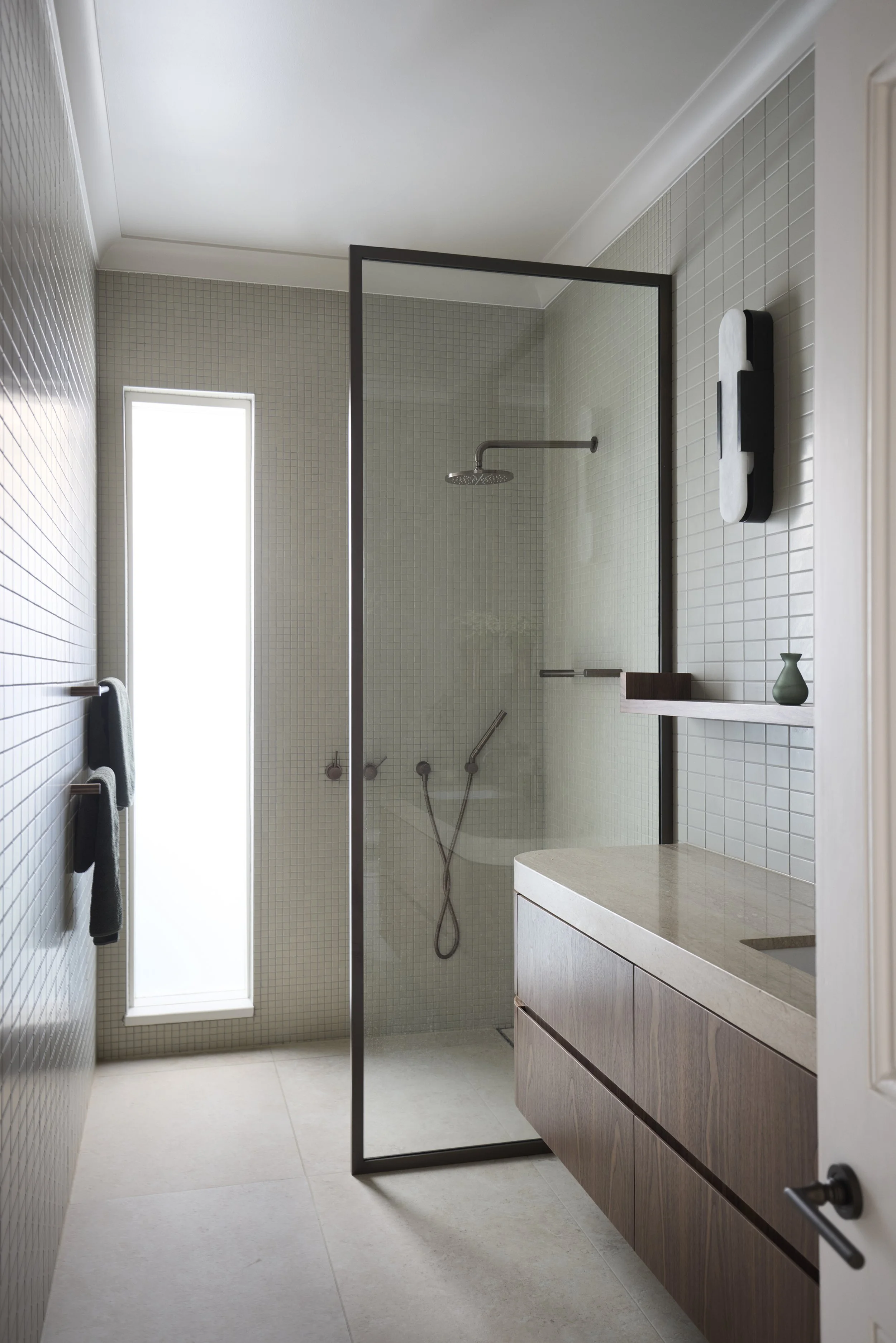 Modern bathroom with glass-enclosed shower, wooden vanity, beige tiled floor, and minimal decor.