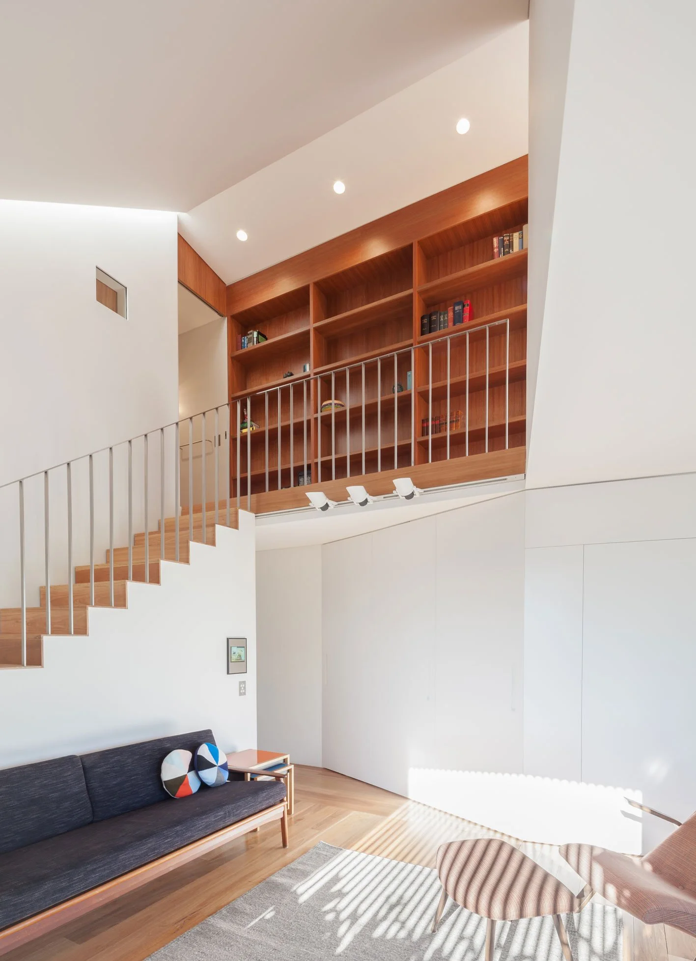 Living room with white walls and wooden flooring, black sofa with decorative pillows, round striped coffee table, and a wooden bookshelf on the upper level with books, overlooking the space below.