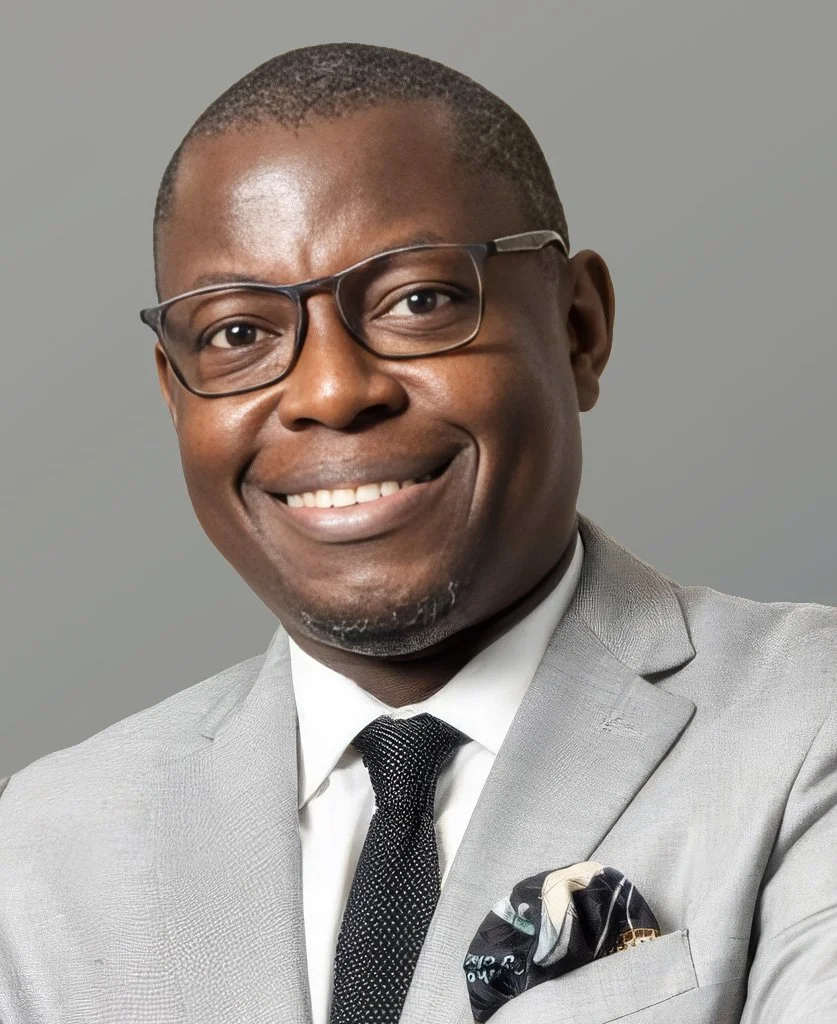 Portrait of an African American man in a light gray suit, white shirt, and black tie, wearing glasses, smiling, against a plain gray background.