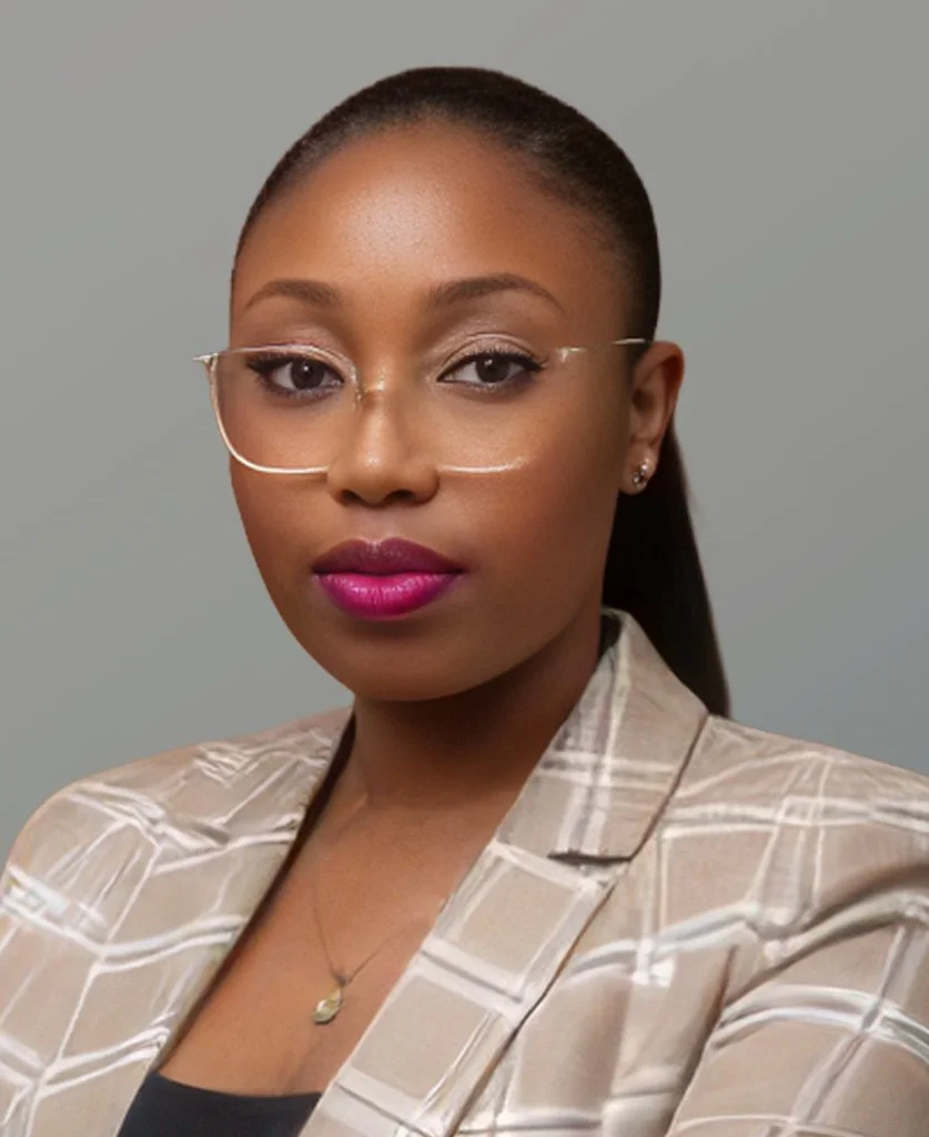 A woman with dark skin, glasses, and makeup, dressed in a beige plaid blazer, accessorized with earrings and a necklace, against a neutral gray background.
