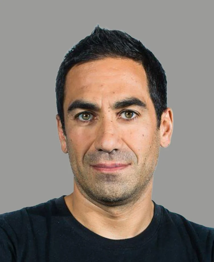 A smiling young man in a navy suit and white shirt against a plain gray background.
