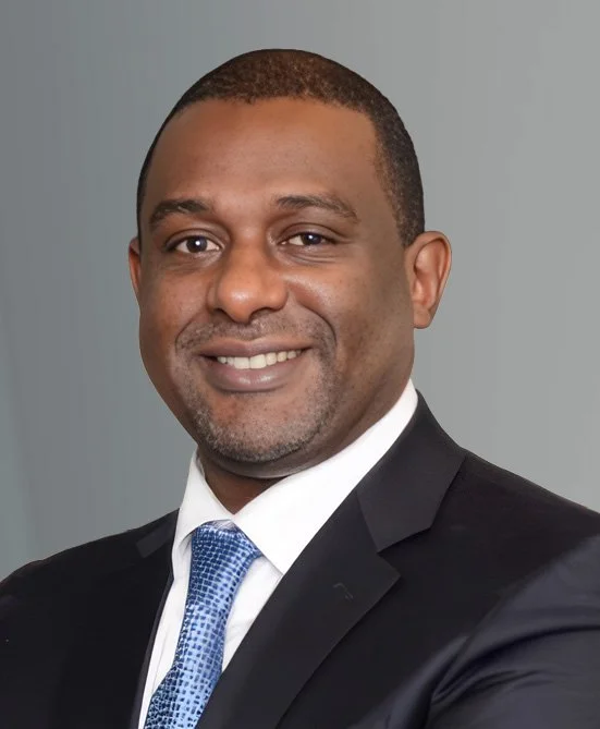 Professional headshot of a man in a suit with a blue tie, smiling against a gray background.