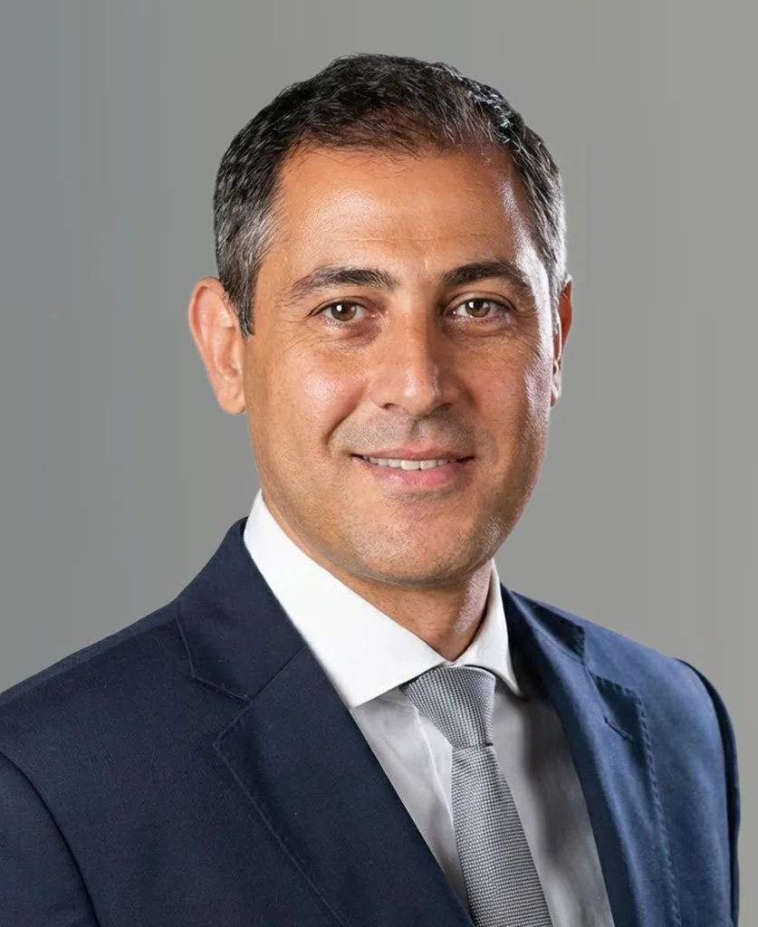 Professional headshot of a man in a navy suit, white shirt, and grey tie, smiling against a neutral grey background.