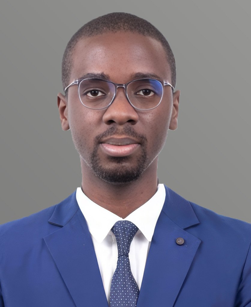 A professional portrait of a young African American man wearing glasses, a white shirt, a blue suit, and a blue dotted tie, against a plain grey background.