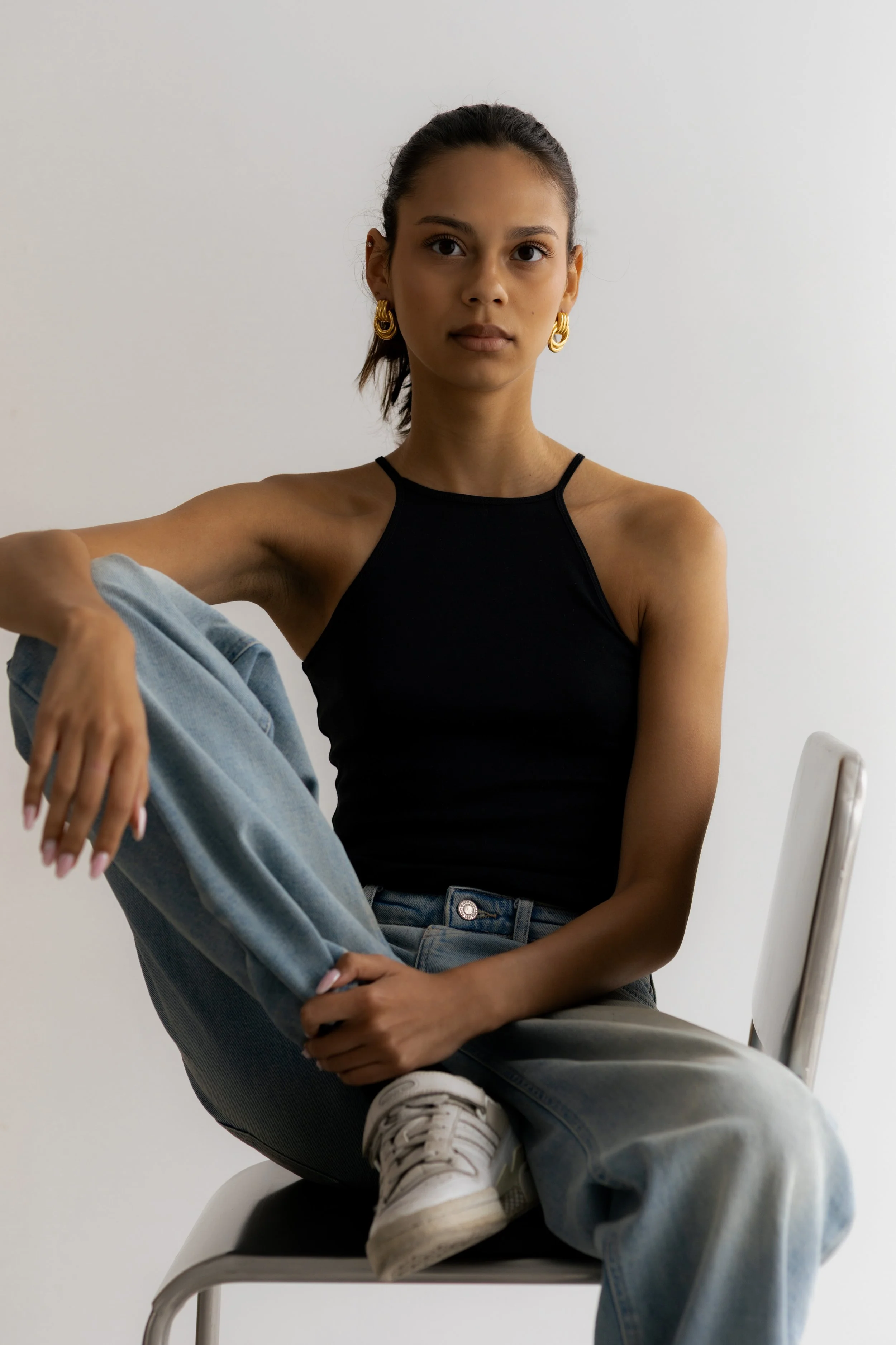A young woman with dark hair tied back, wearing a black tank top, gold hoop earrings, and light blue jeans, seated on a white chair against a plain white background.