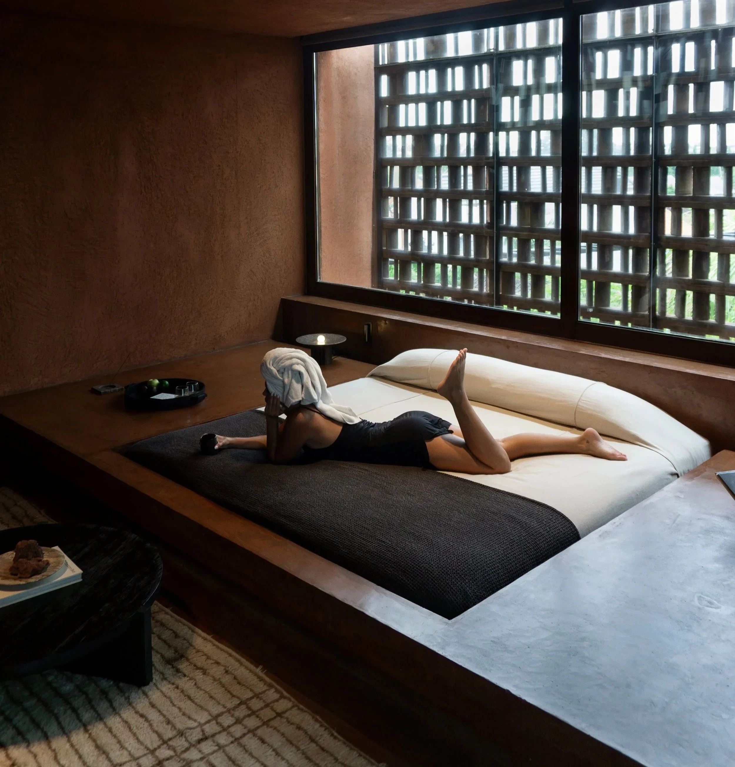 Person lying on a bed with a towel on their head, inside a room with a large window and wooden accents.