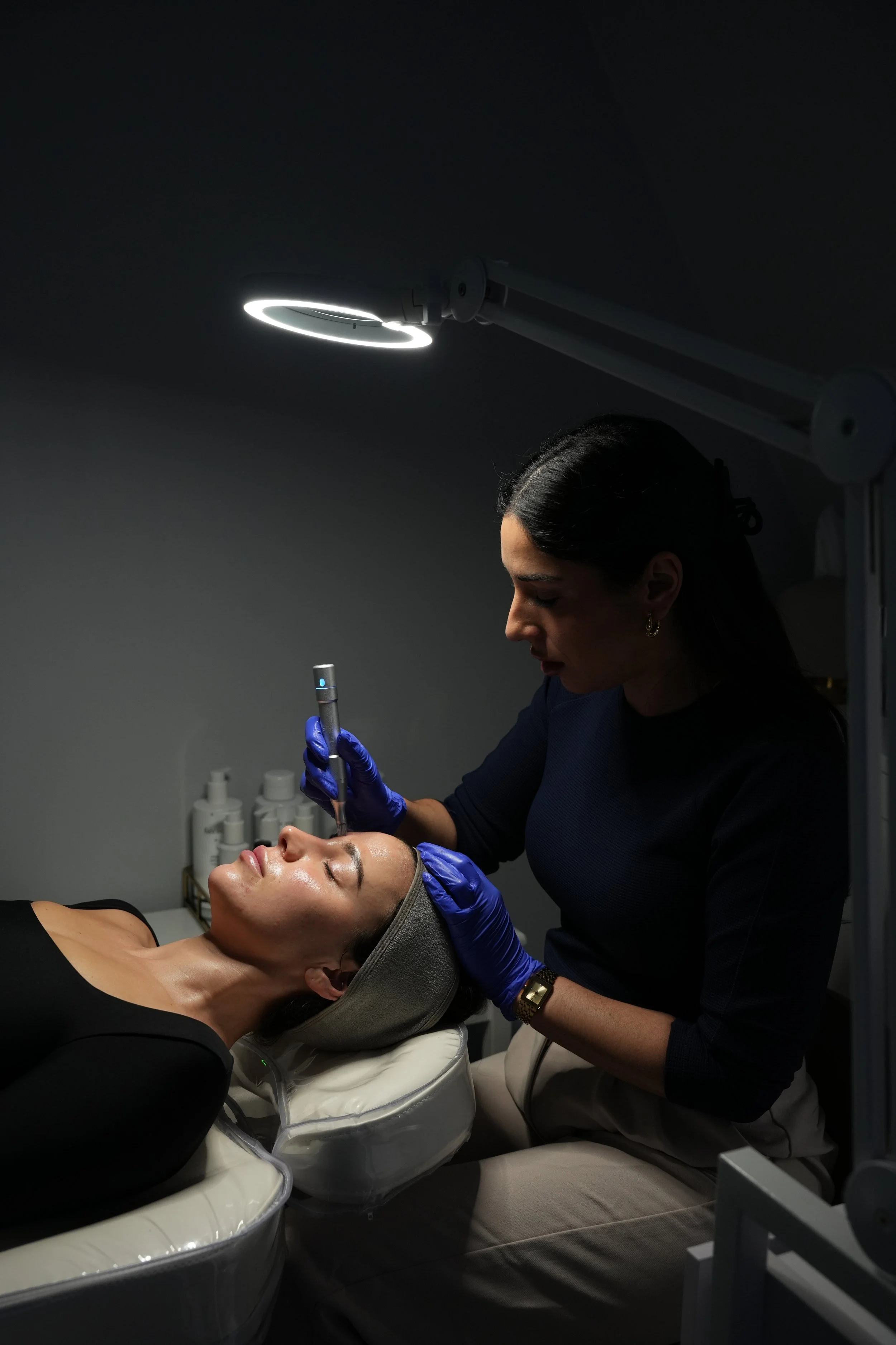 A woman receiving a cosmetic treatment on her face from a professional in a dimly lit clinic.