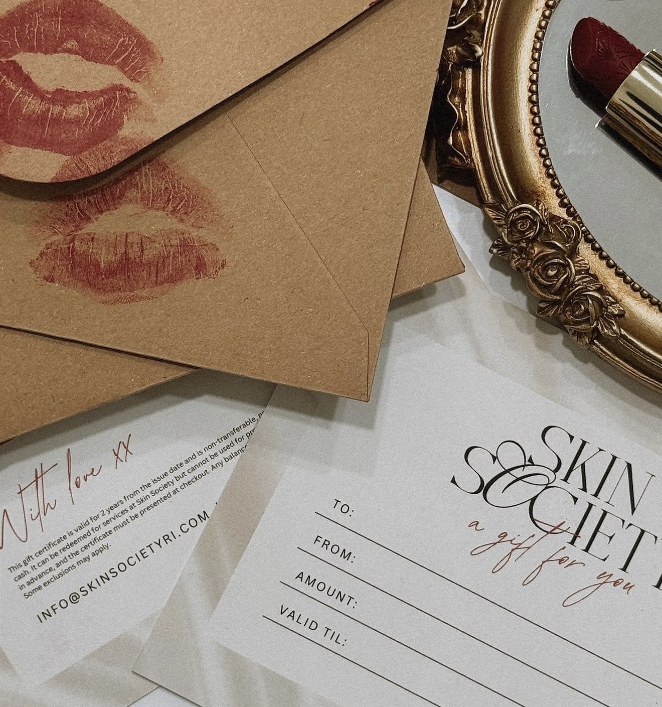 Gift card with a lipstick and a makeup mirror on a white surface, envelopes with kissing lips prints, and a gift card with blank fields for recipient, sender, amount, and expiry date.