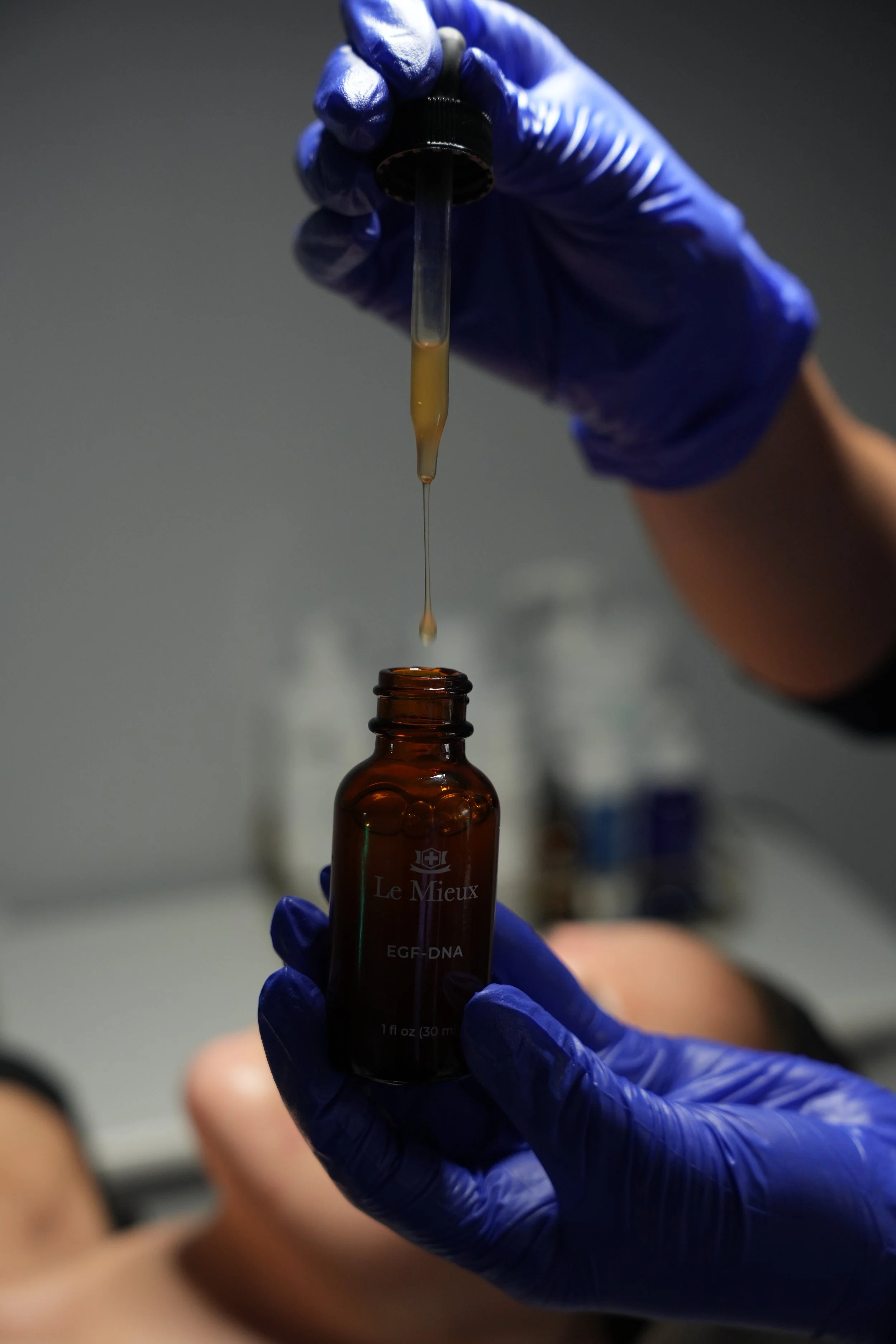 Person wearing blue gloves handling a small dropper with a yellowish liquid over a brown glass bottle labeled 'Le Mieux' and 'EGF-DNA'.