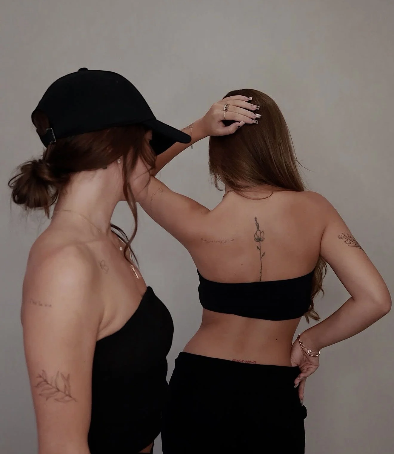 Two women with tattoos posing against a plain background, one wearing a black cap and the other with a strapless top.