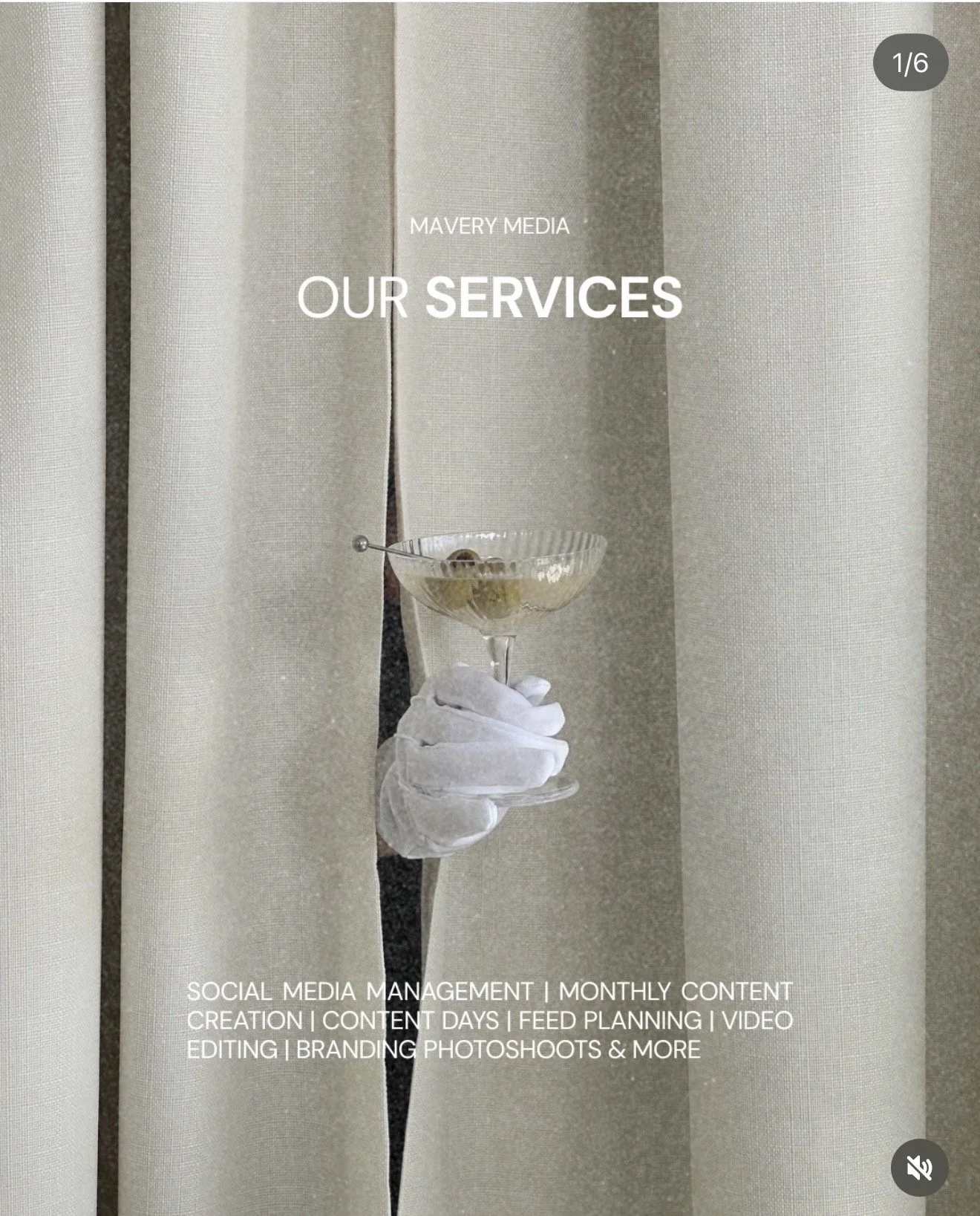 A person wearing white gloves holding a cocktail glass with green olives, partially hidden behind closed beige curtains.