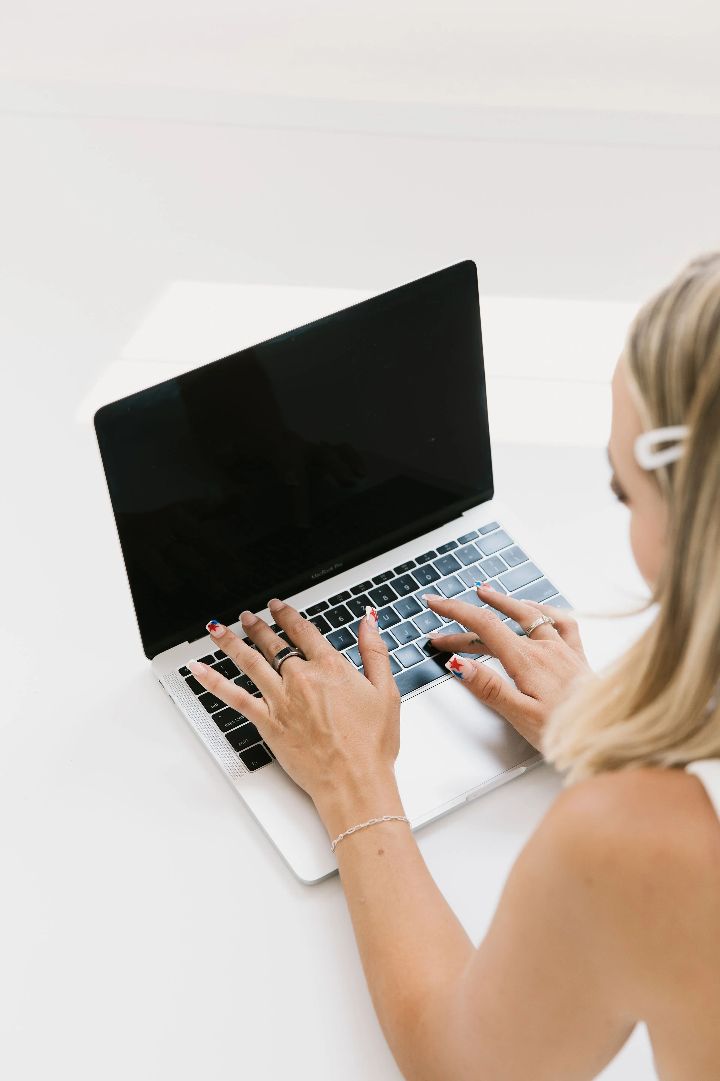 A woman with blonde hair, wearing hair clips, rings, and a bracelet, is using a laptop with a black screen on a white desk.