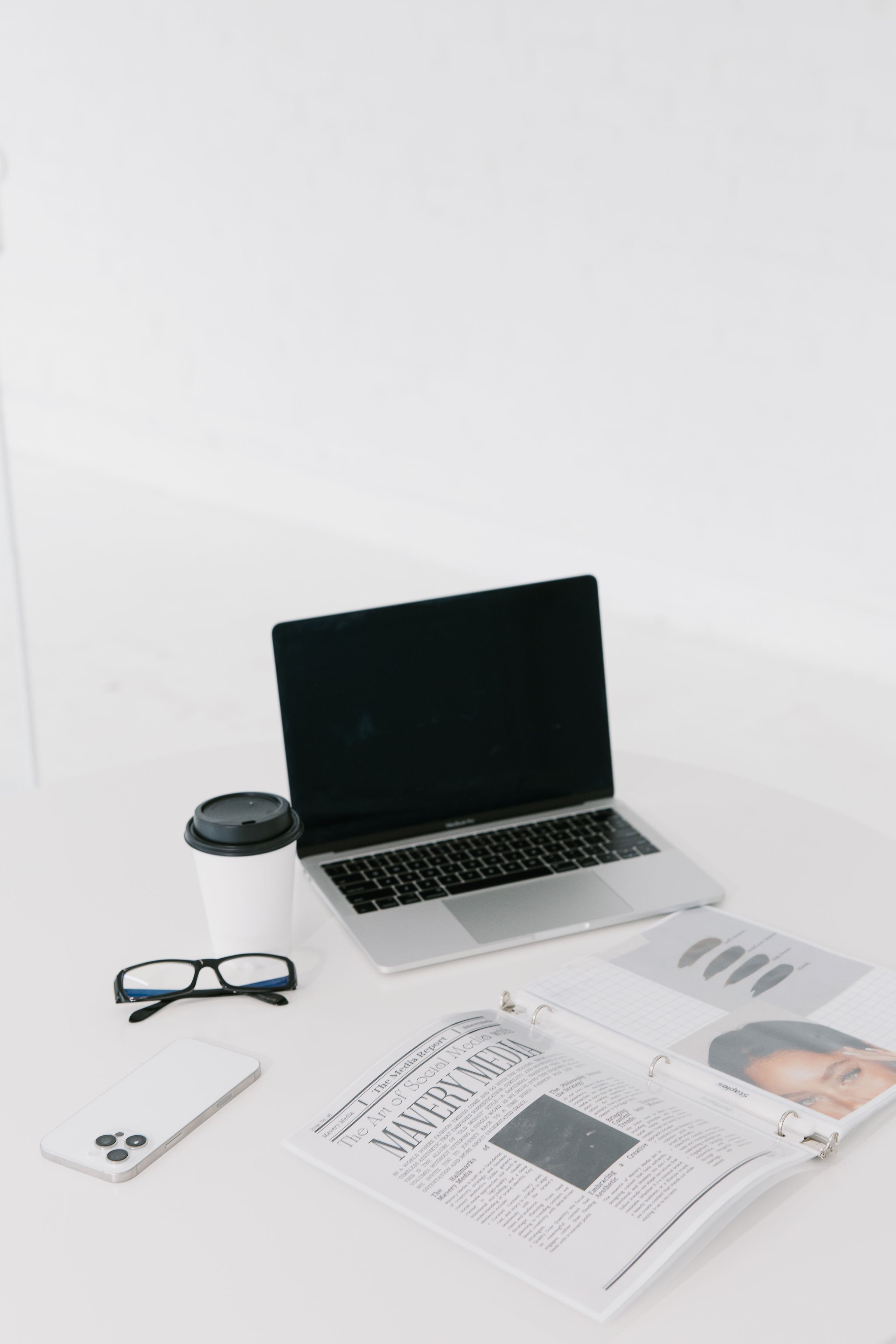 A minimalist white desk with a laptop, a disposable coffee cup, a pair of black glasses, a white smartphone, and an open magazine with articles and images.
